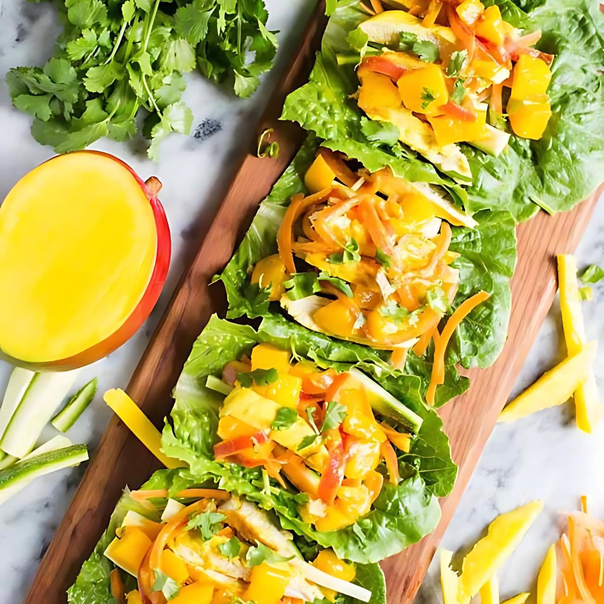  Several mango chicken lettuce wraps arranged on a wooden board. The wraps contain shredded chicken, mango, and a sweet and savory sauce, garnished with cilantro. A half of a mango, cucumber slices, and cilantro sprigs are also on the board.