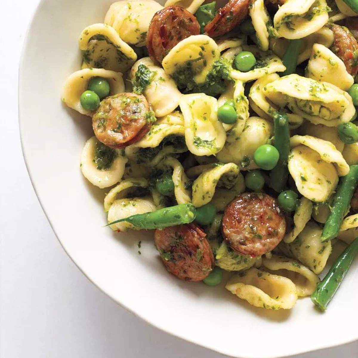 A bowl of orecchiette pasta tossed with pesto, cooked sausage, green peas, and snow peas, served on a white plate.