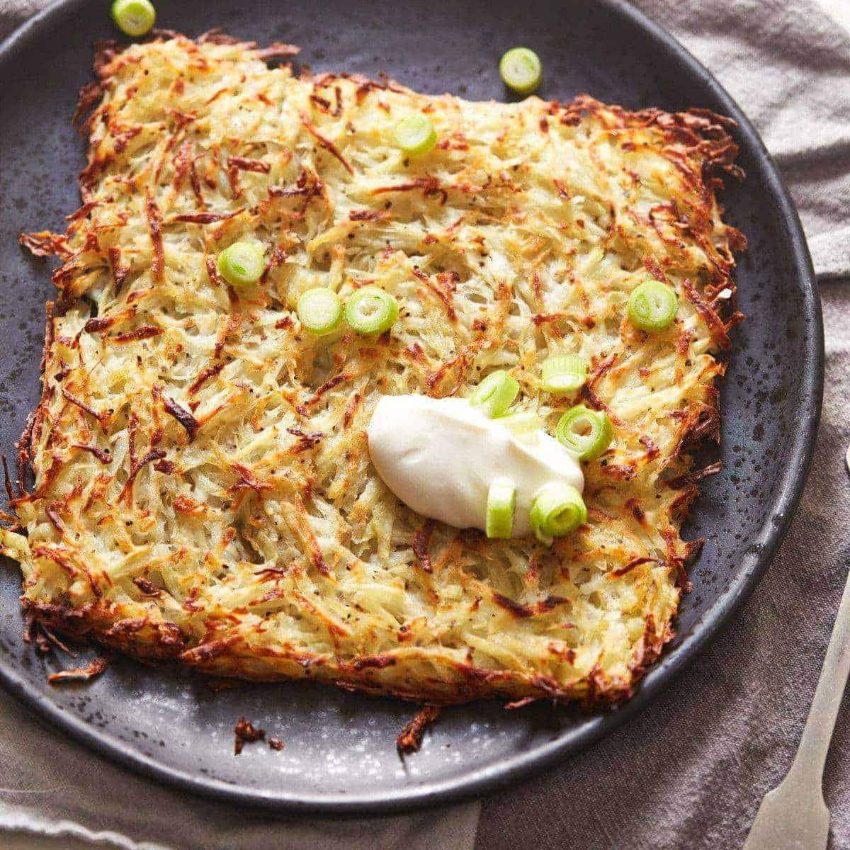  A crispy potato rosti topped with sour cream and chopped green onions served on a black plate.