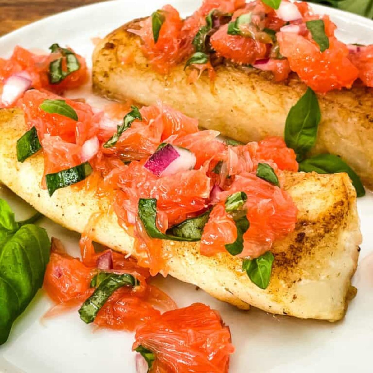  Two pan-seared fish fillets topped with a grapefruit relish and garnished with fresh basil served on a white plate.