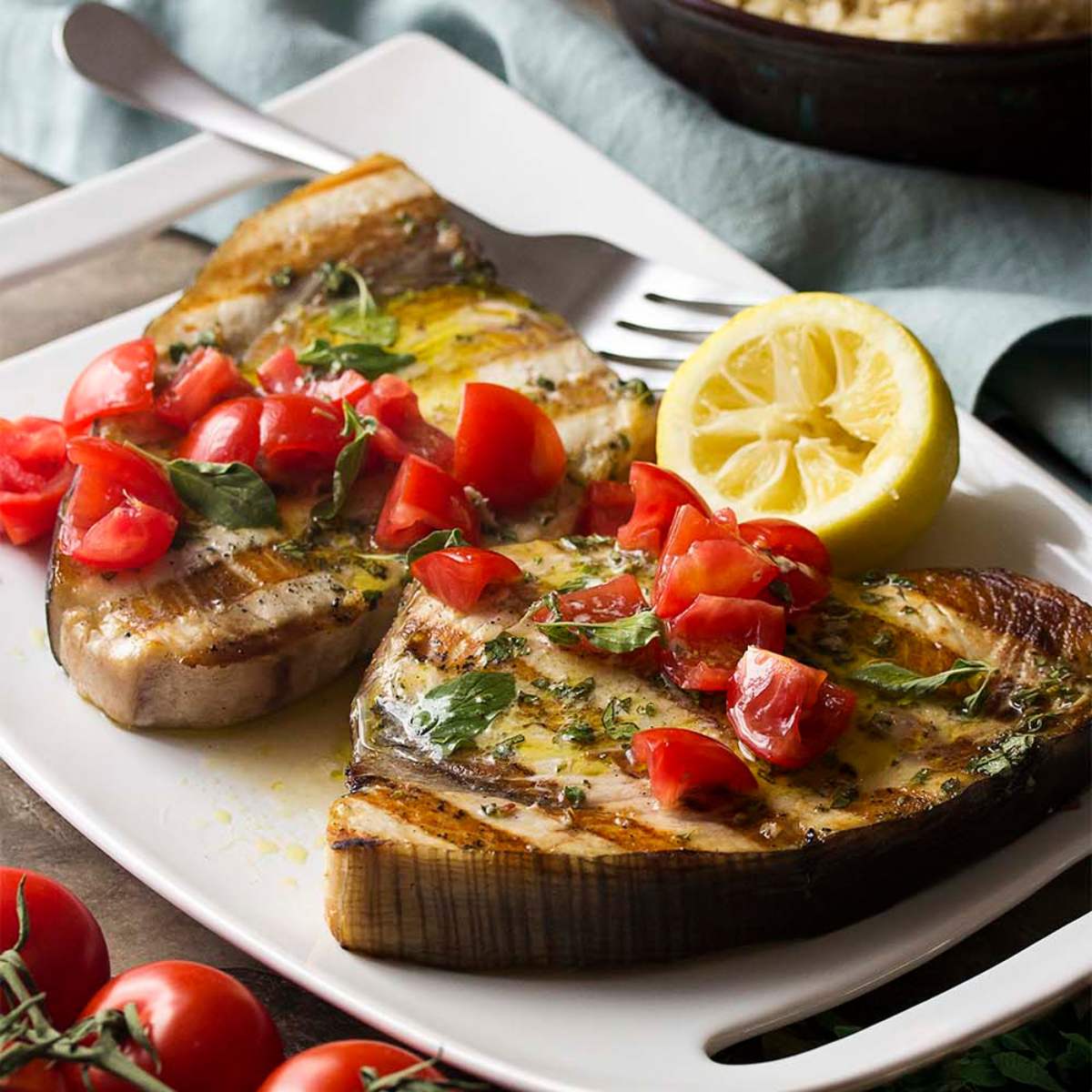 Grilled fish steaks topped with a tomato, basil salsa, and lemon wedge served on white plate. There are cherry tomatoes in the background as well.