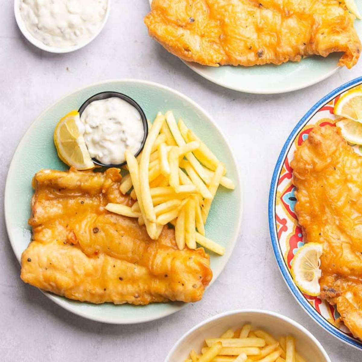 Fish and chips served on plates with tartar sauce, lemon wedges, and a side of fries.

