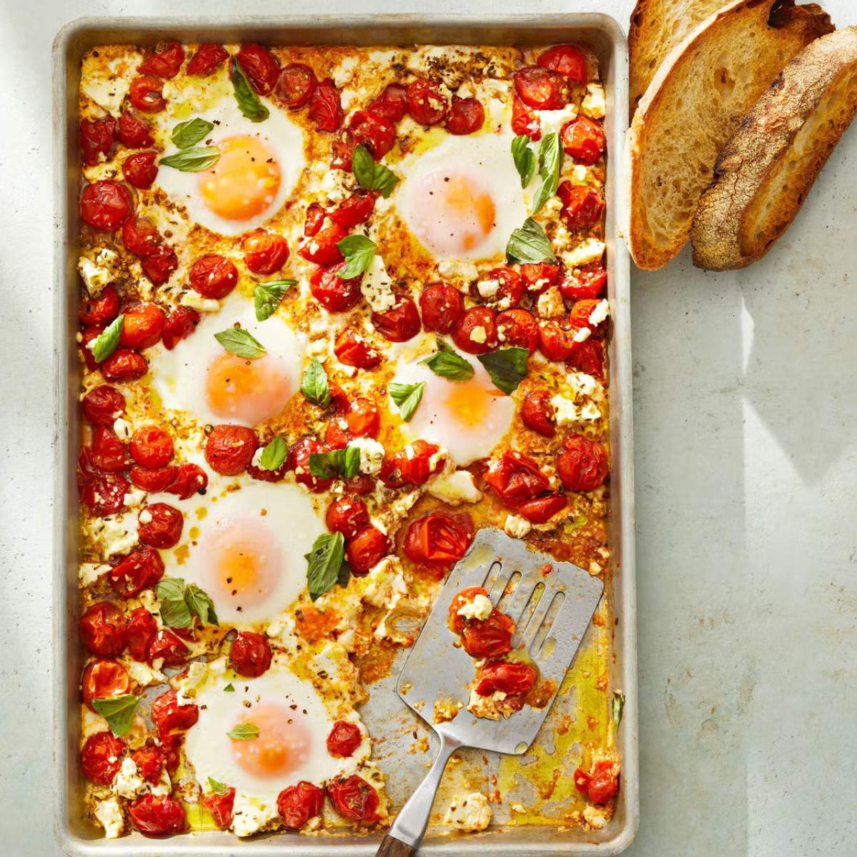  Baked eggs with tomatoes, feta cheese, and basil, served on a baking sheet with toasted bread on the side.
