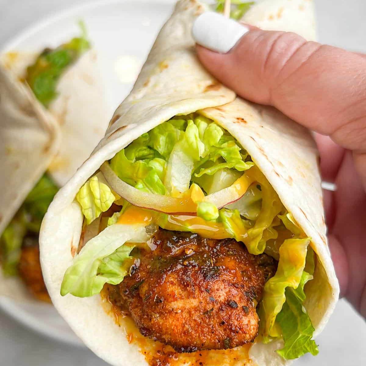 A photo of a chicken wrap being held in someone's hand. The wrap is filled with shredded blackened chicken, lettuce, and a yellow sauce.