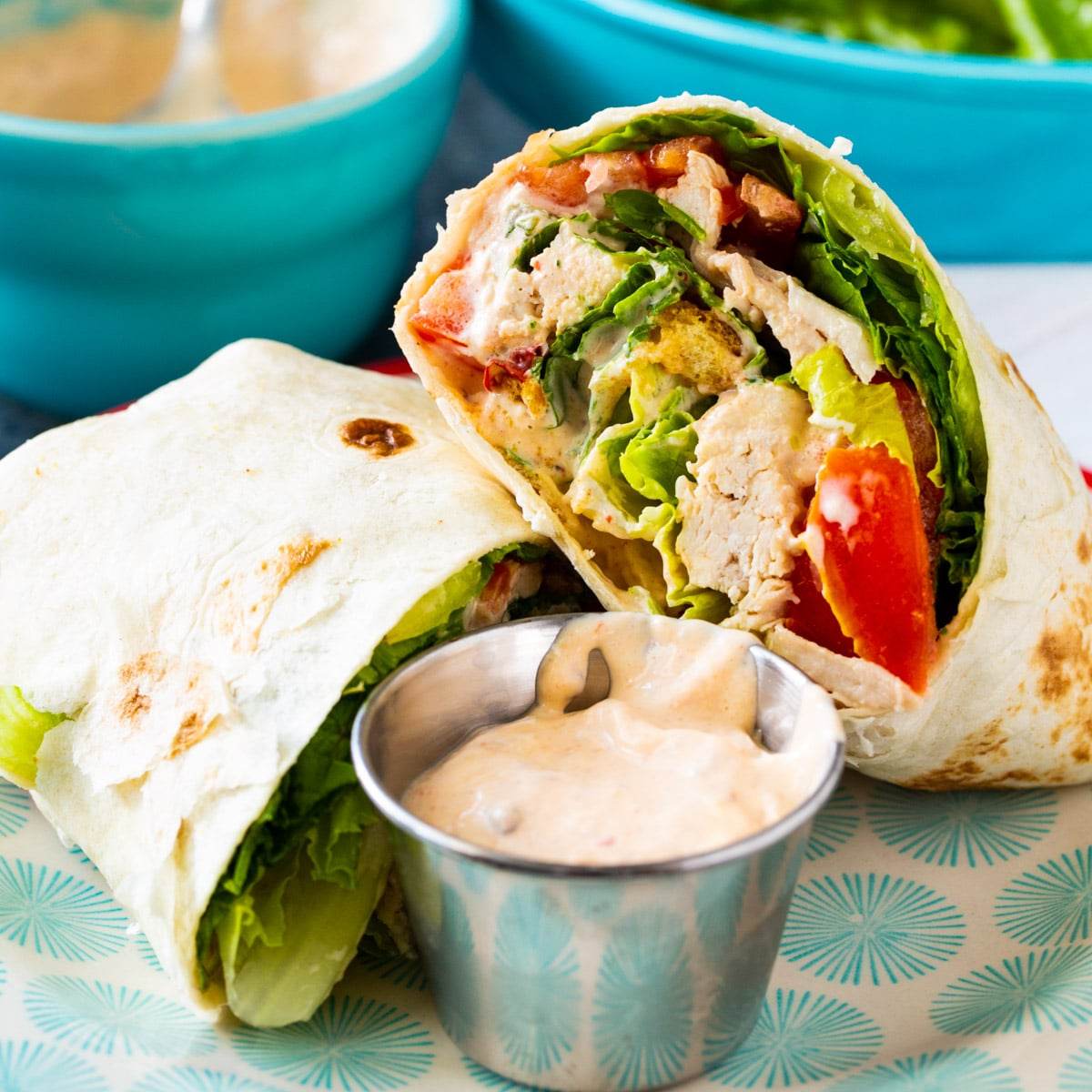 Chicken Caesar Salad Wrap cut in half, showing grilled chicken, romaine lettuce, tomatoes, and a creamy Caesar dressing inside a tortilla. A small bowl of ranch dressing is also on the plate.