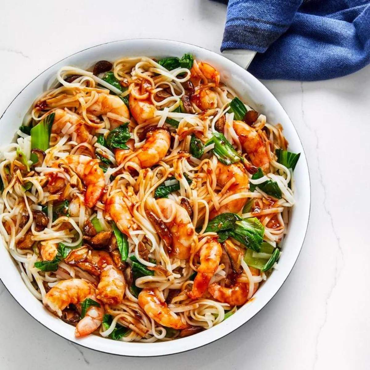  A white bowl filled with stir-fried rice noodles. The noodles are coated in a glossy, reddish-brown sauce and are mixed with cooked shrimp, chopped vegetables.