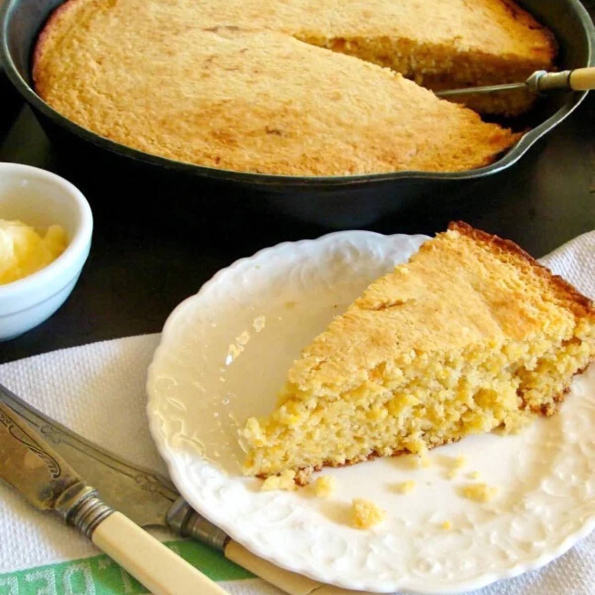 A golden-brown cornbread loaf in a cast iron skillet, with a slice on a plate next to it. A small bowl of butter and a knife and fork are also visible.