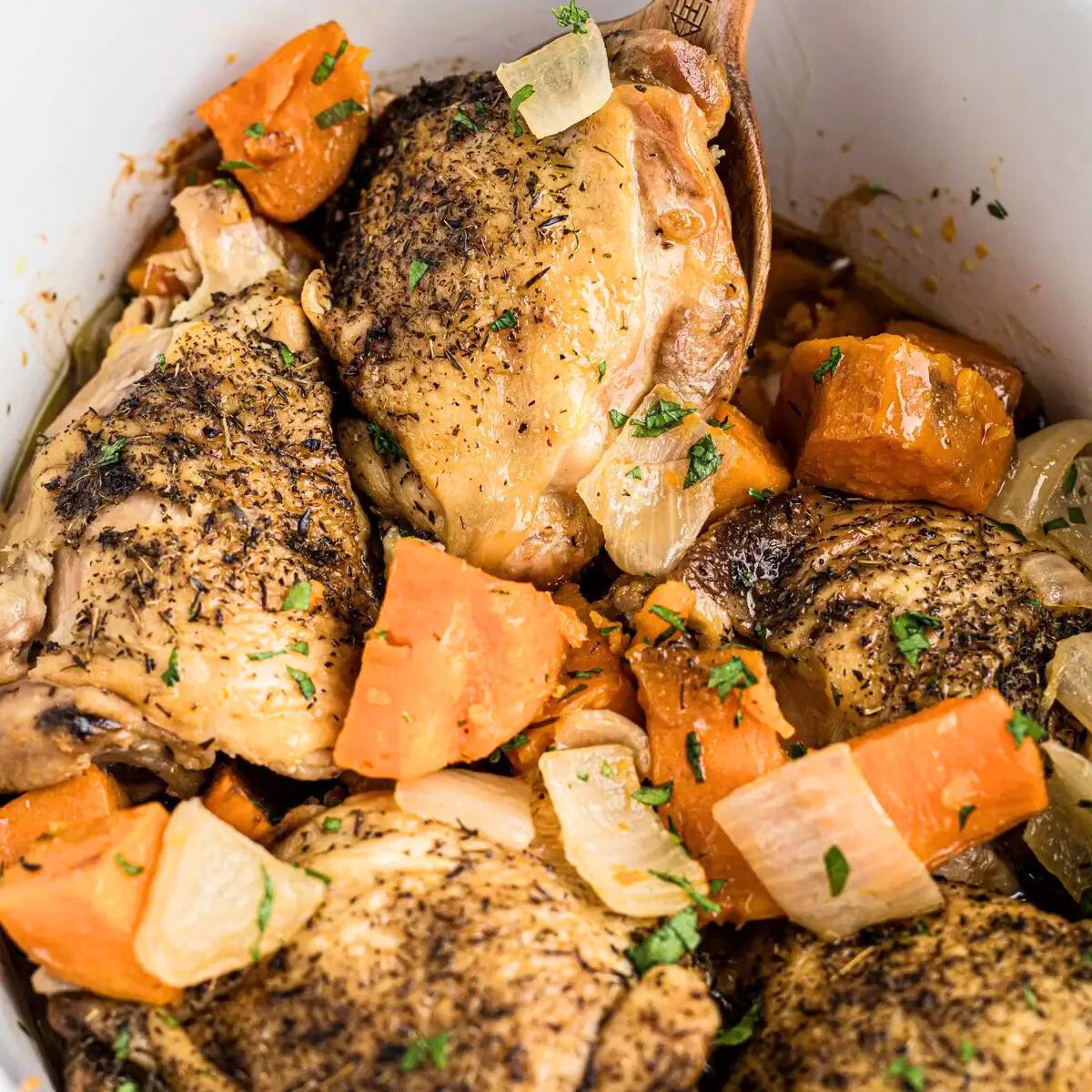 A slow cooker filled with chicken thighs, sweet potatoes, carrots, and onions. The chicken is seasoned with herbs and spices, and the vegetables are coated in a flavorful broth.