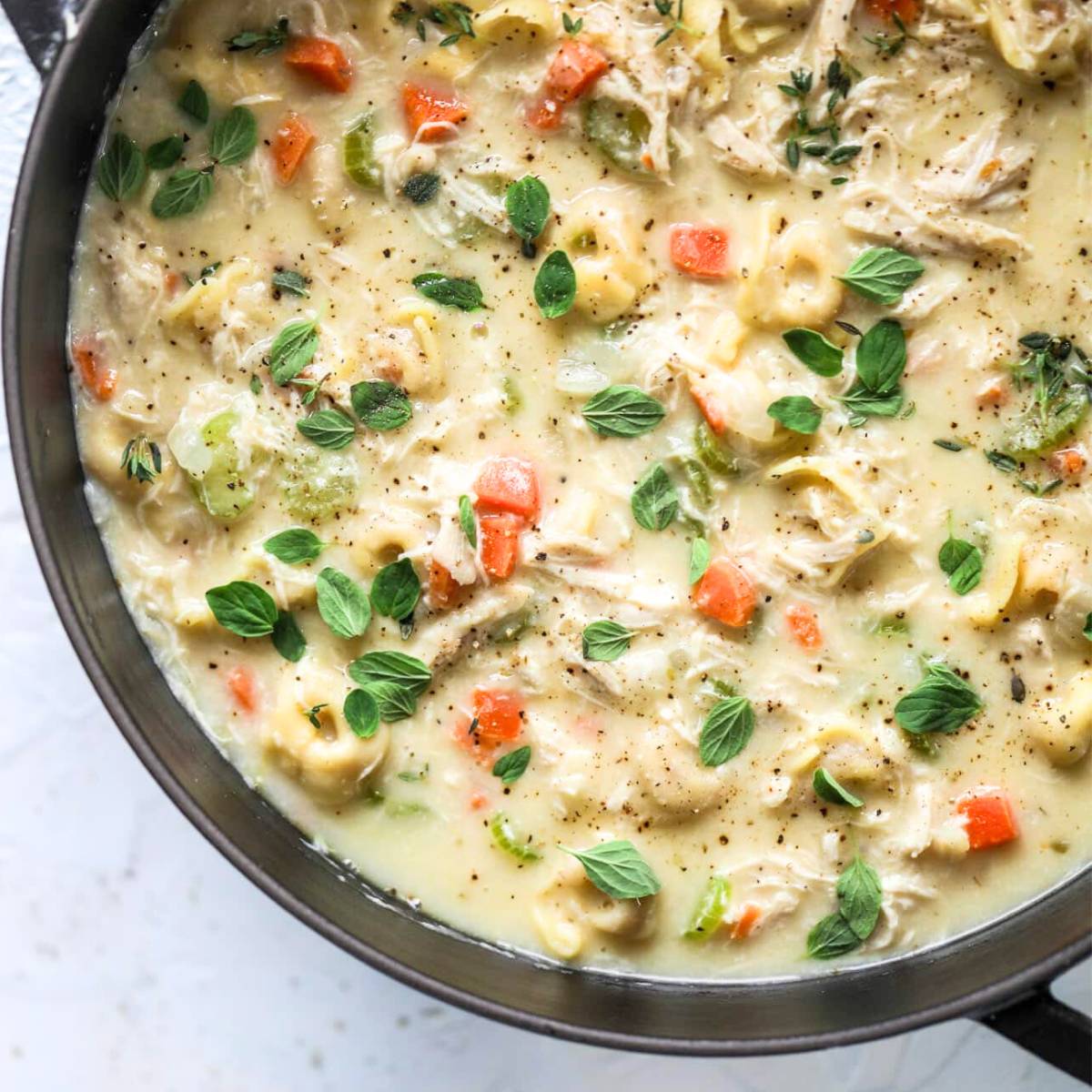 A large pot filled with creamy chicken tortellini soup. The soup contains visible tortellini pasta, shredded chicken, carrots, and celery. It is topped with fresh thyme leaves and cracked black pepper.