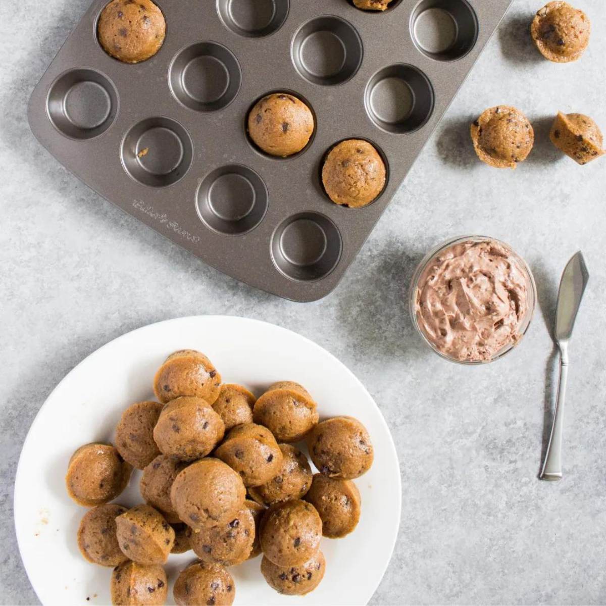  muffin tin with some mini muffins already baked, a pile of baked mini muffins on a plate, a small bowl of mocha cream cheese spread, and a knife on a gray surface.