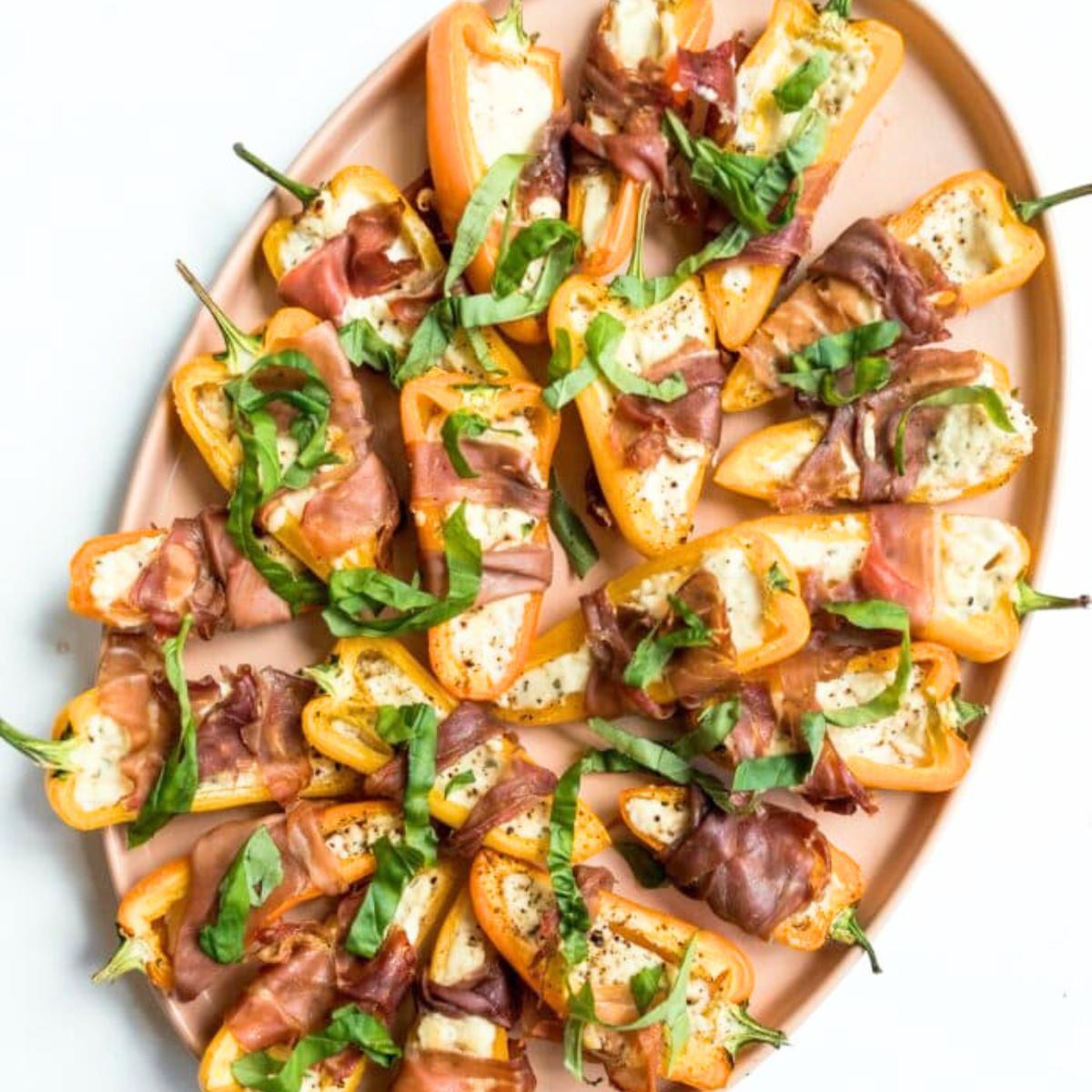  Prosciutto-wrapped mini peppers filled with a creamy cheese mixture, garnished with fresh basil. Served on a platter.