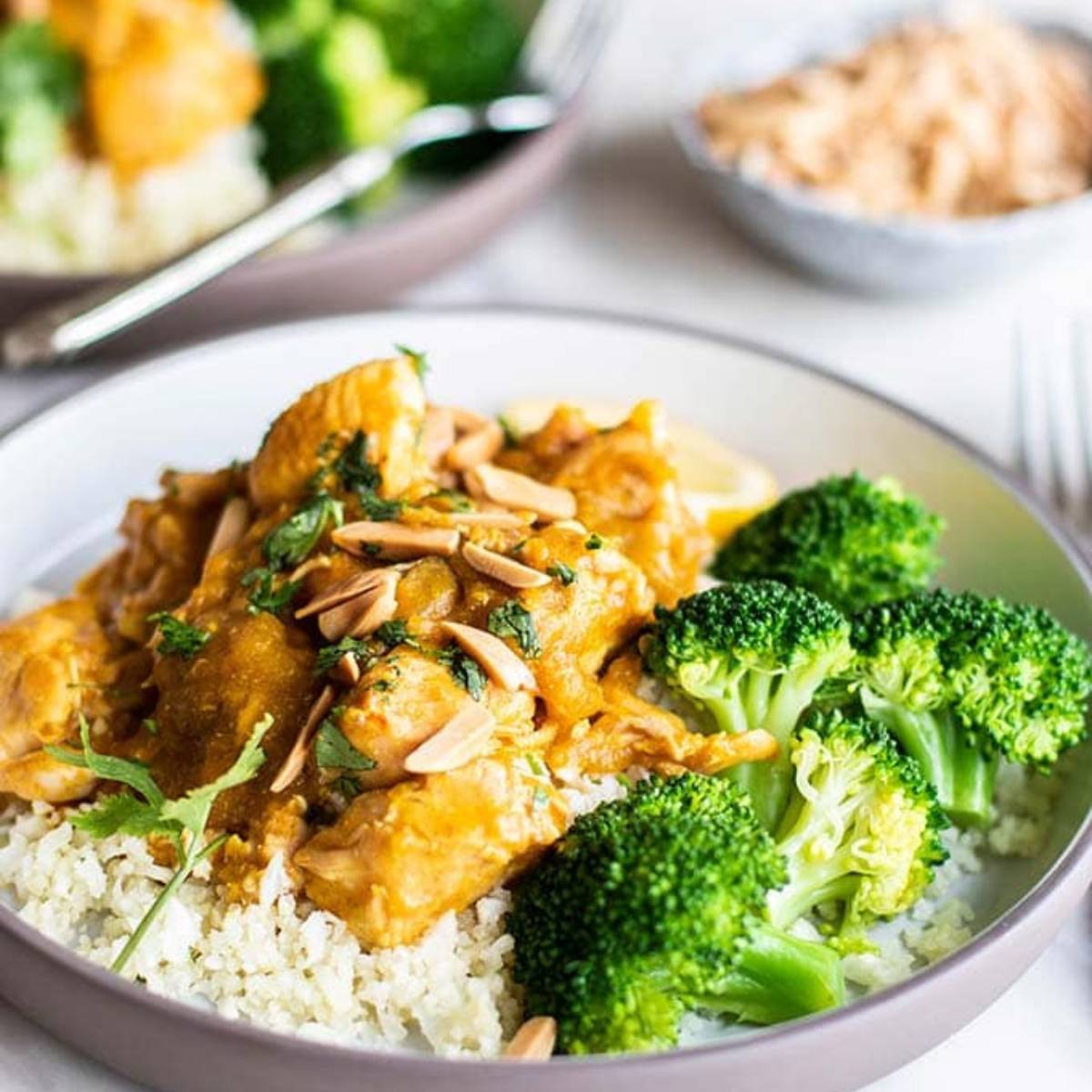 A plate of creamy coconut curry chicken served over cauliflower rice. The chicken is coated in a flavorful curry sauce and garnished with almonds and fresh cilantro. A side of steamed broccoli is served alongside.