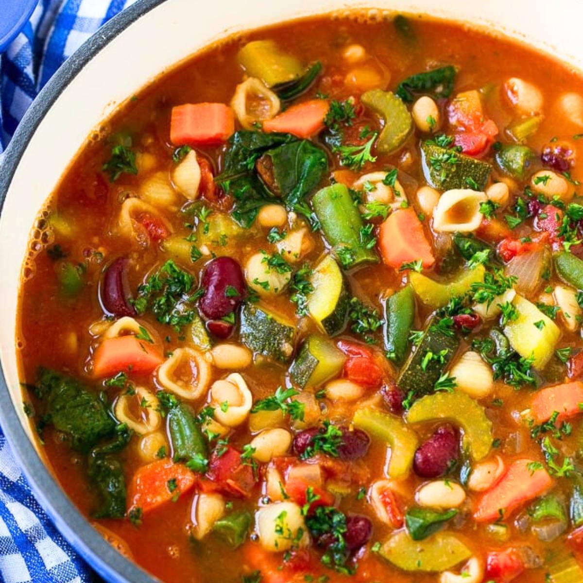 A large pot filled with a hearty minestrone soup, packed with vegetables like carrots, zucchini, beans, and spinach. Small pasta shells are also visible throughout the soup, which is topped with fresh parsley.