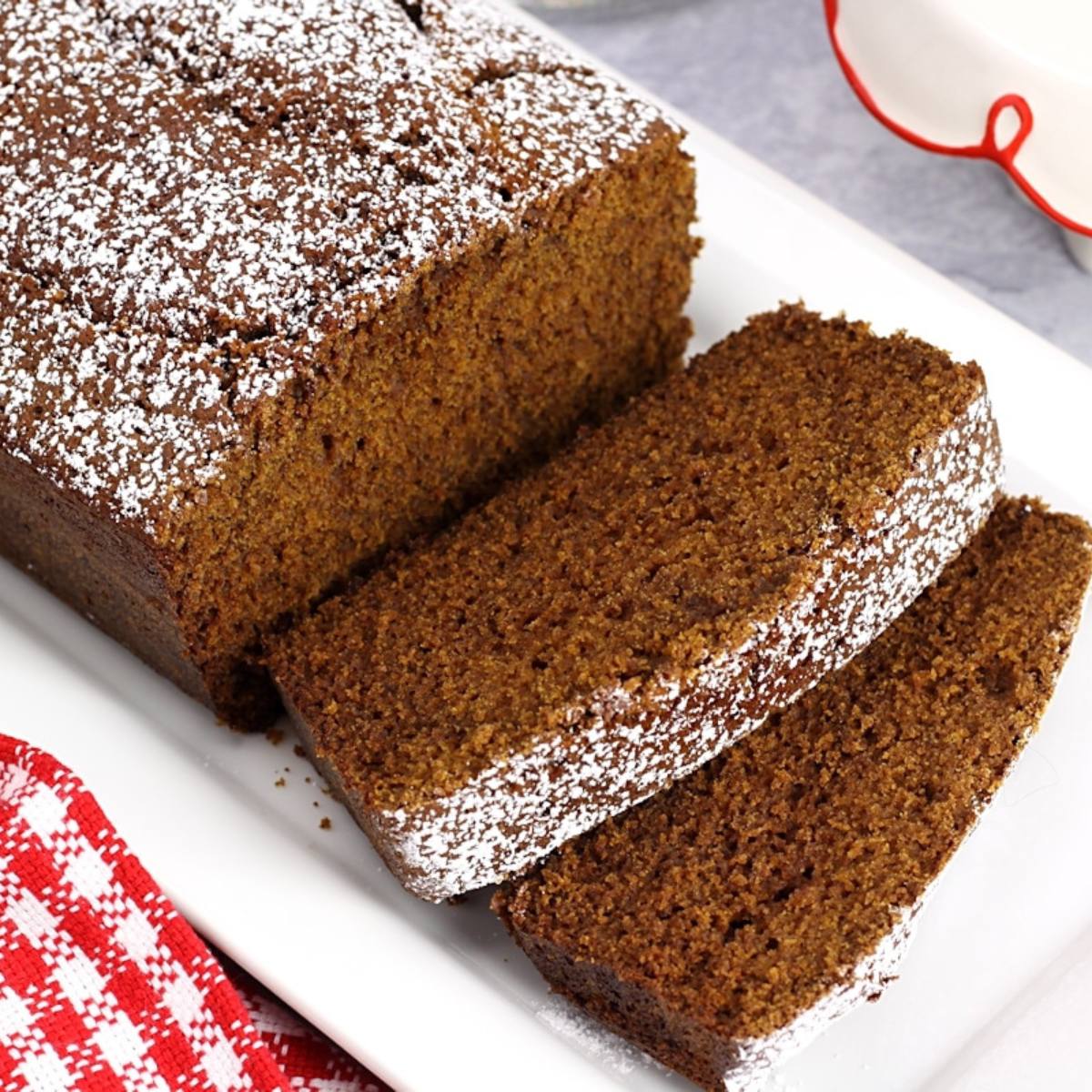 A loaf of gingerbread bread, sliced and dusted with powdered sugar, on a white plate.