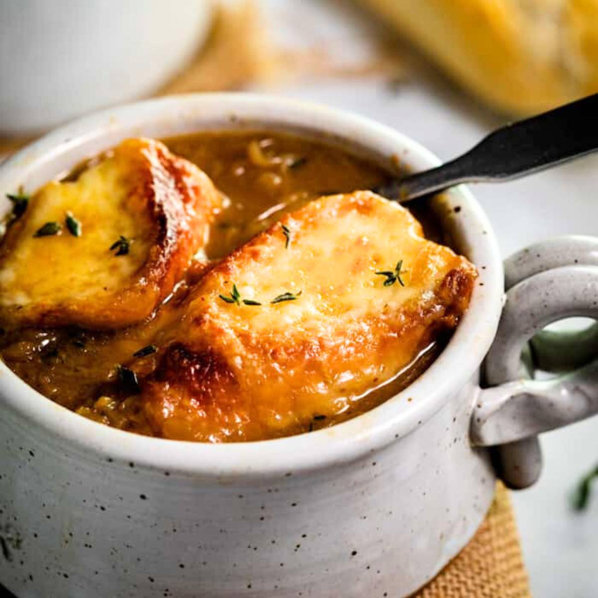  A bowl of French onion soup with a golden brown crouton topped with melted cheese and fresh thyme. The soup is a rich, brown color and is served in a white bowl.