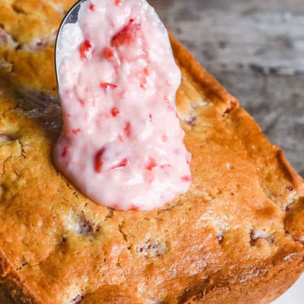 A spoon drizzling pink strawberry glaze onto a loaf of golden-brown bread.