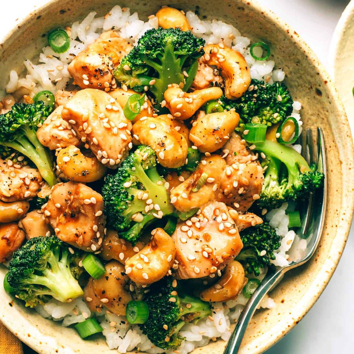 A bowl of cashew chicken and broccoli served over rice. The dish is topped with sesame seeds and chopped green onions.