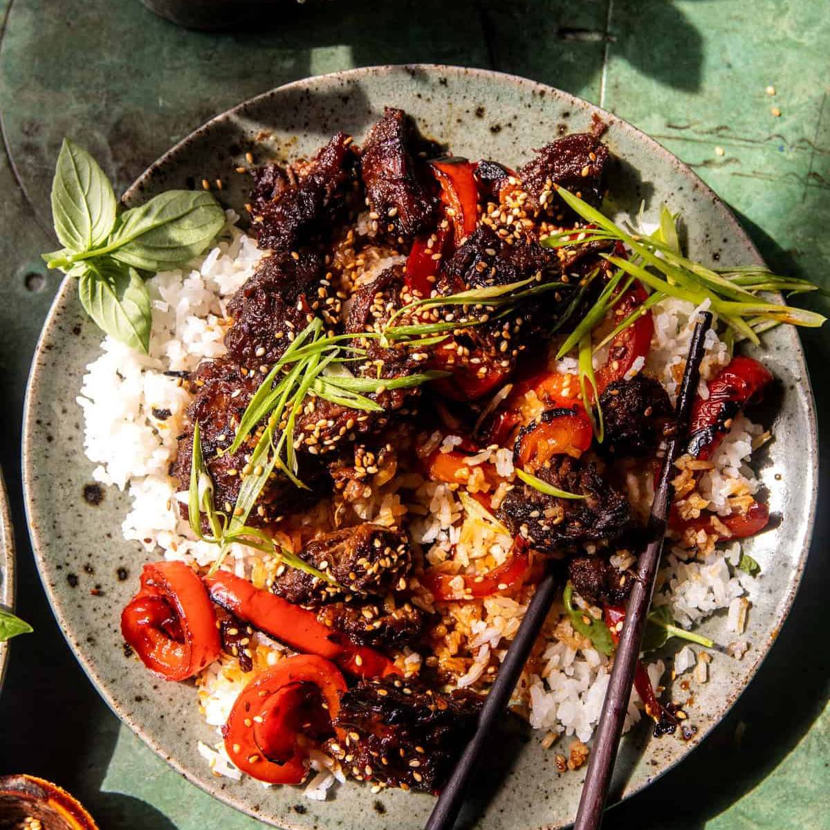 A plate of Korean-style beef bulgogi served over white rice. The beef is stir-fried with bell peppers and coated in a sweet and savory sauce. It's garnished with sesame seeds and chopped green onions. 