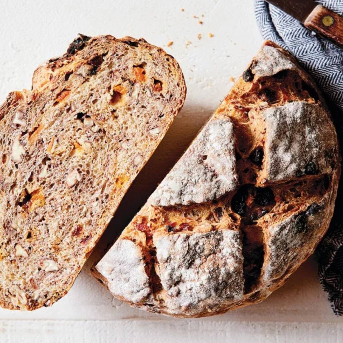 A loaf of dark, crusty bread with dried fruit and nuts, sliced in half on a white surface.