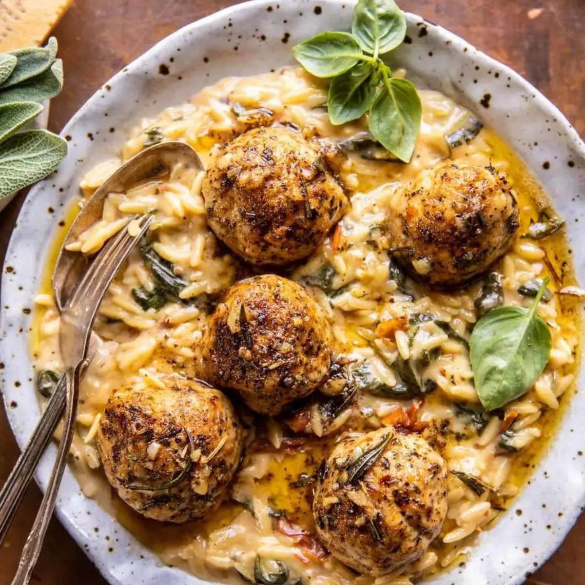 A bowl of creamy chicken meatballs served with orzo pasta, spinach, and Parmesan cheese. The dish is garnished with fresh basil leaves.