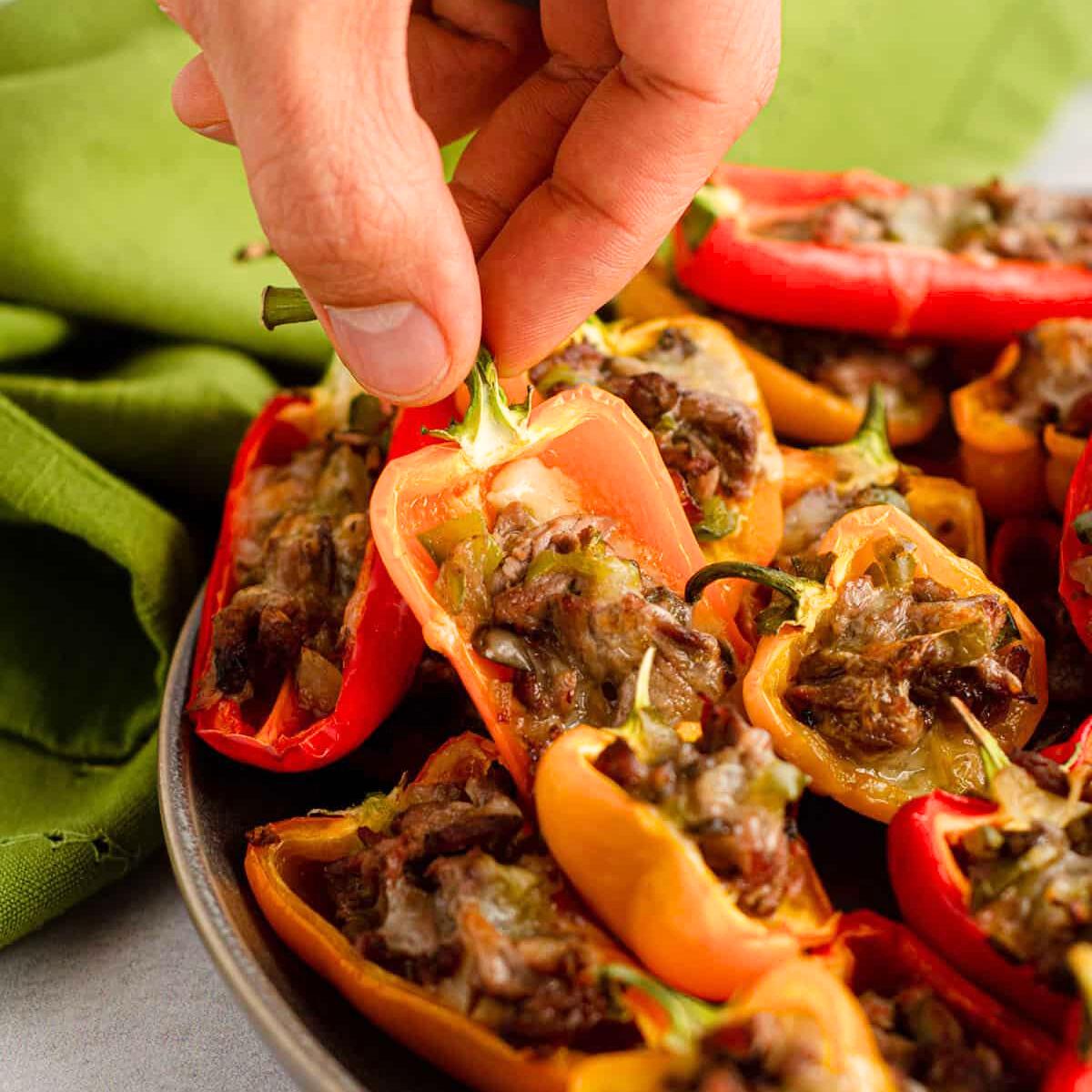 A hand picking up a stuffed mini pepper from a plate of other stuffed mini peppers. The peppers are filled with a meat and cheese mixture.