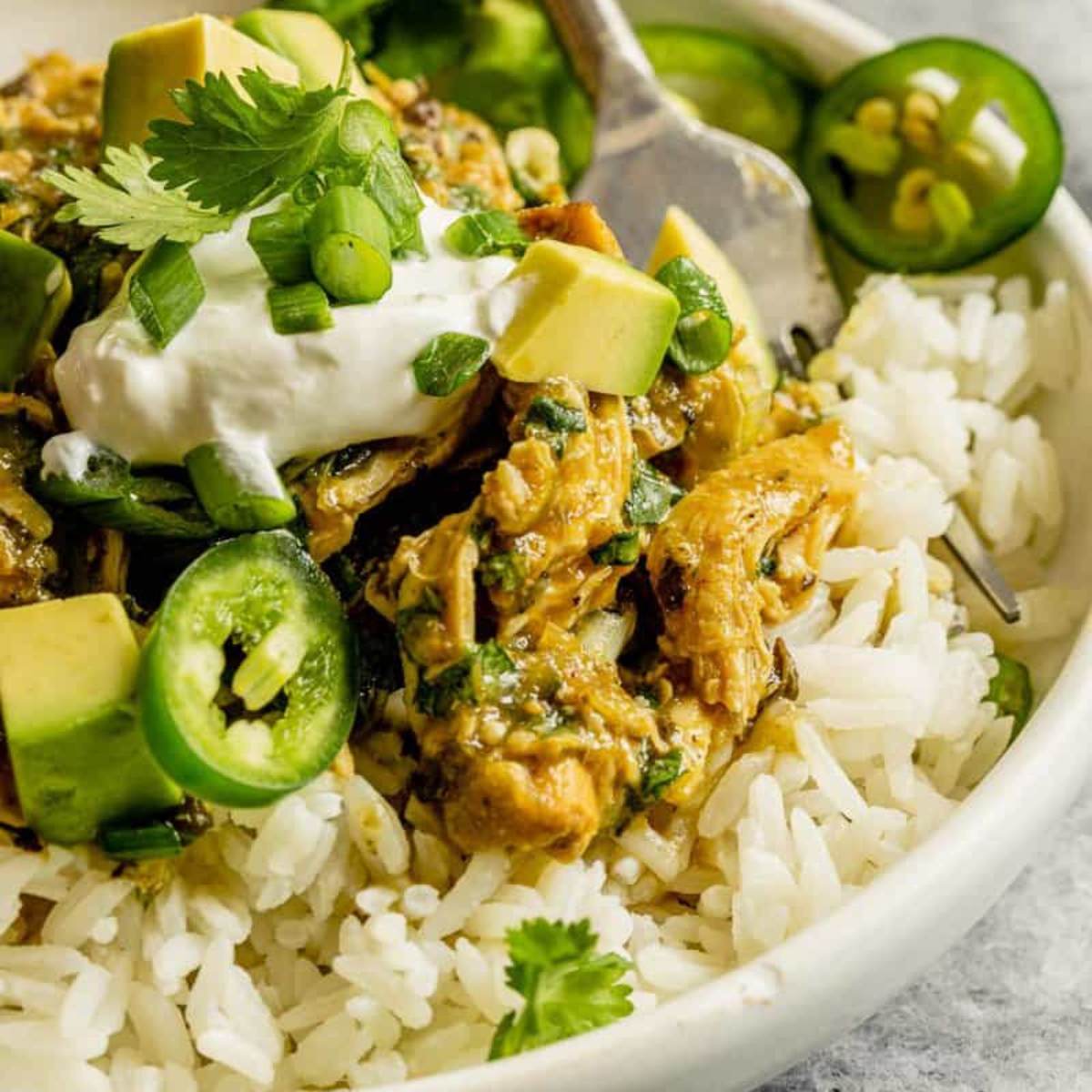  A bowl of Salsa Verde Chicken served over white rice. The chicken is topped with chopped avocado, sour cream, and sliced jalapeño peppers.