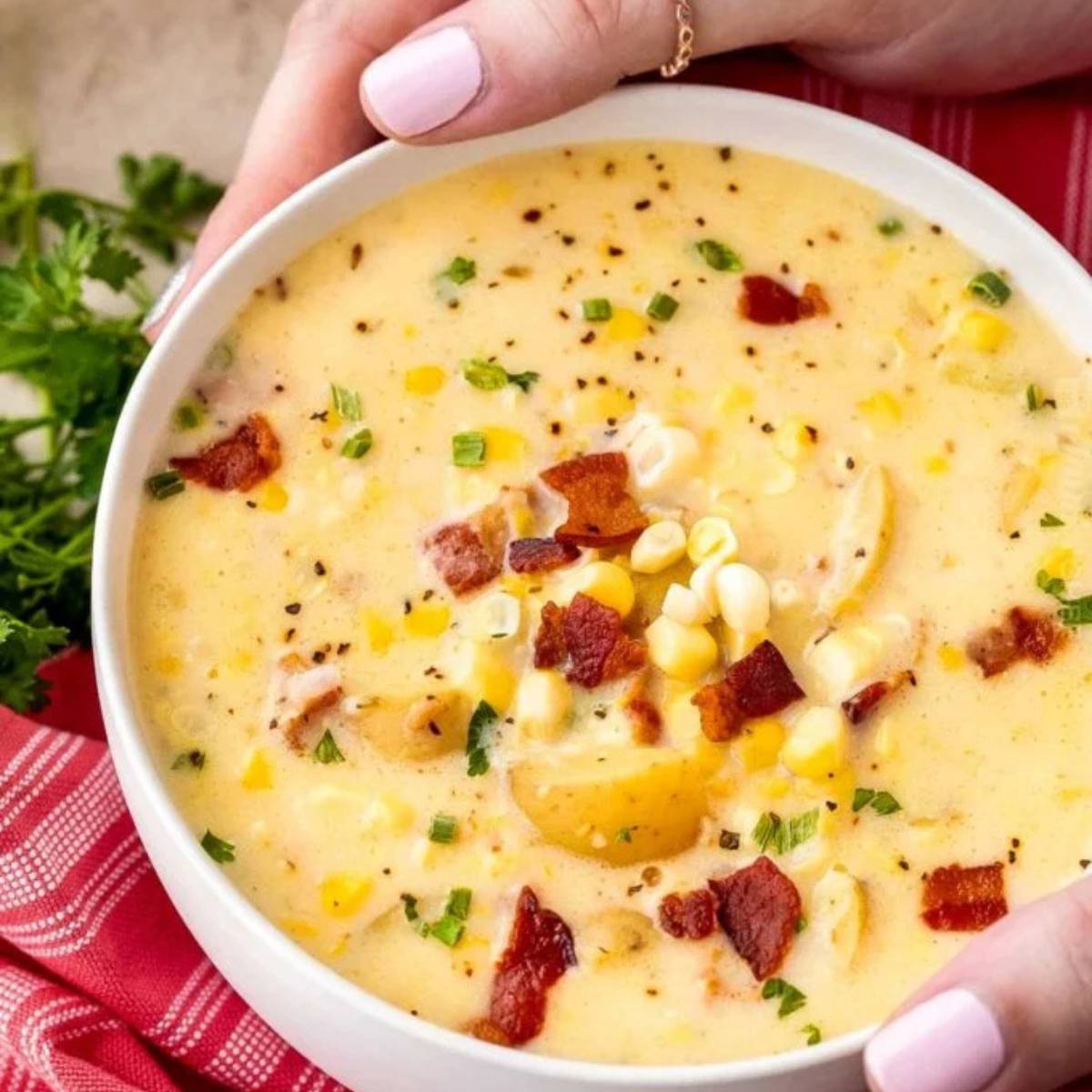 A bowl of creamy corn chowder, topped with crumbled bacon, chopped green onions, and a sprinkle of black pepper. The soup is a pale yellow color and contains visible chunks of corn, potatoes, and bacon.