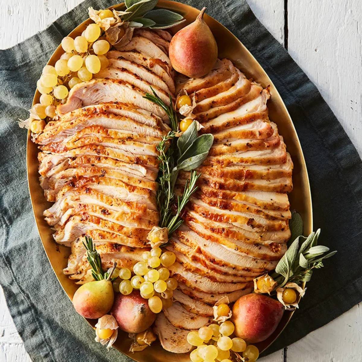 A platter of sliced turkey breast with a golden brown crust. Fresh rosemary and sage sprigs are placed on top, and grapes and pears surround the turkey.