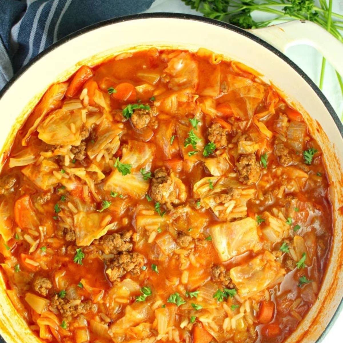 A large pot filled with hearty cabbage roll soup, with visible chunks of ground beef, shredded cabbage, carrots, and rice. The soup is a vibrant red color and is topped with fresh parsley.