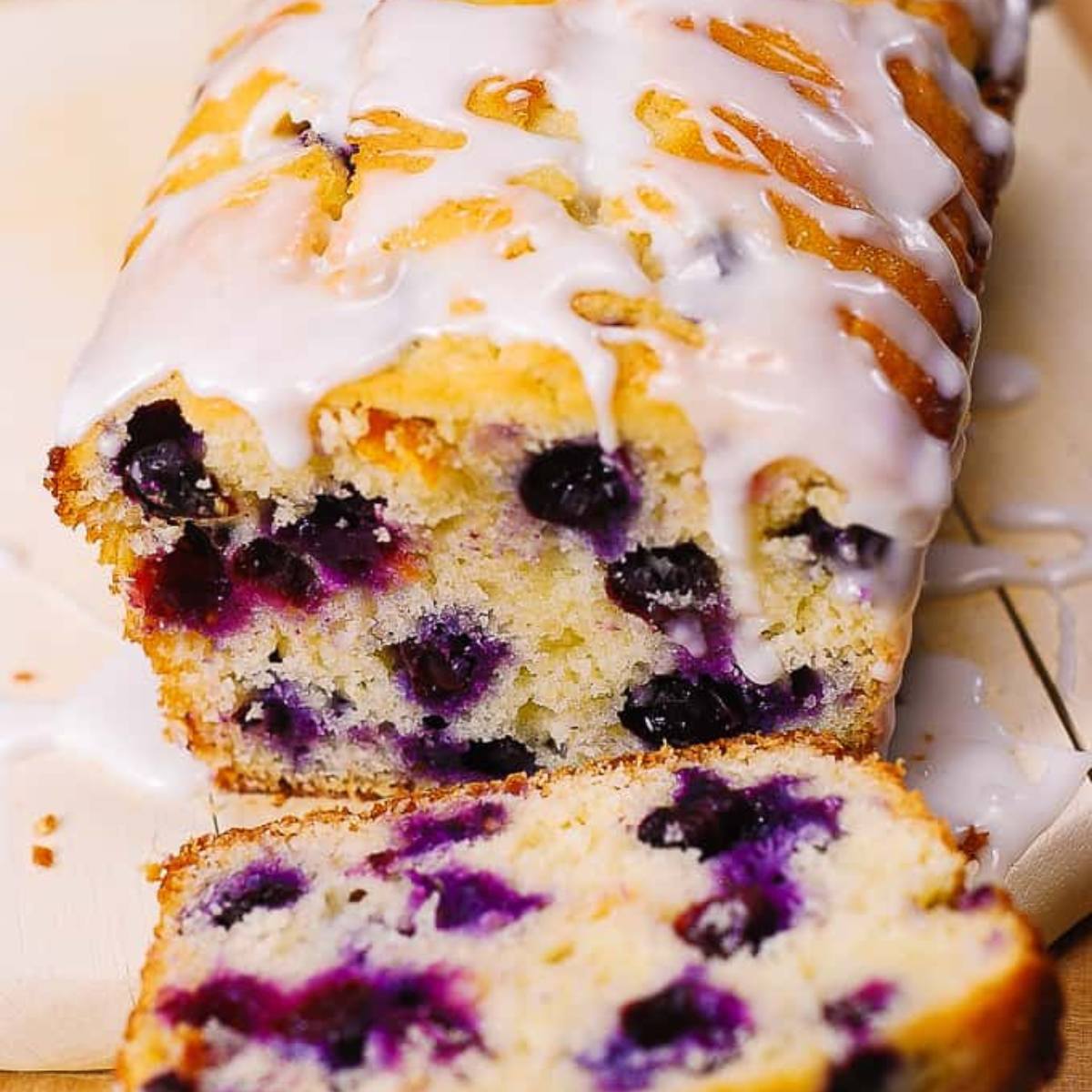 A golden-brown loaf of blueberry bread with a white glaze, partially sliced, on a wooden cutting board.