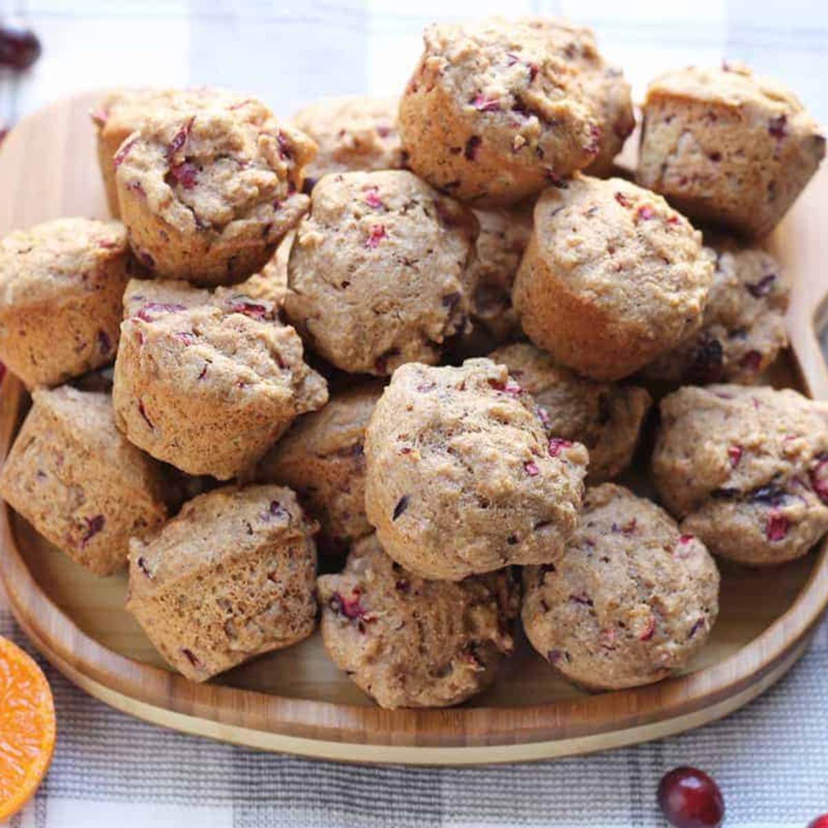  A pile of mini cranberry muffins on a wooden serving tray, with fresh cranberries and oranges in the background.