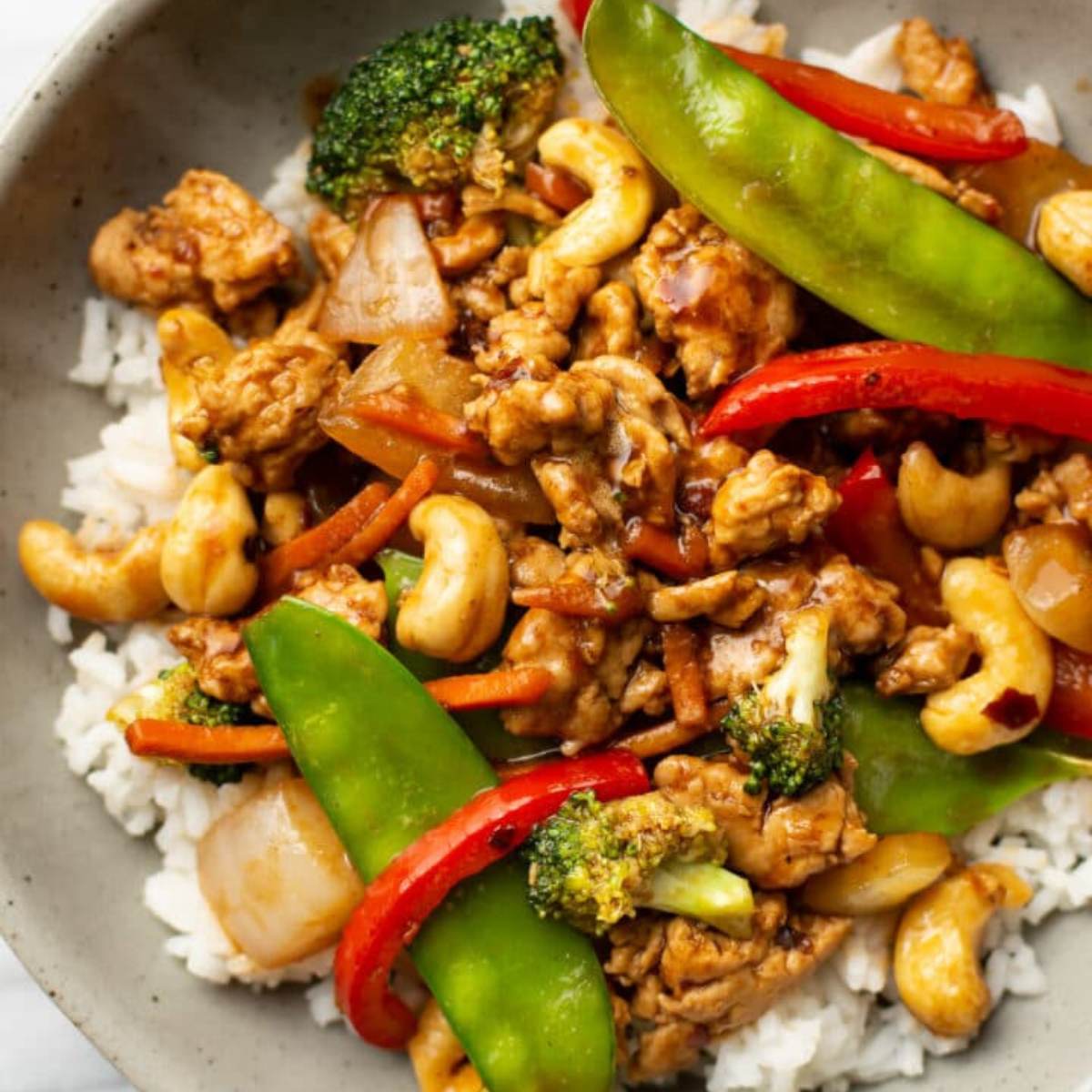 A bowl of cashew chicken stir-fry with rice, featuring chicken, broccoli, carrots, red bell peppers, snow peas, and cashews. The dish is tossed in a savory sauce.