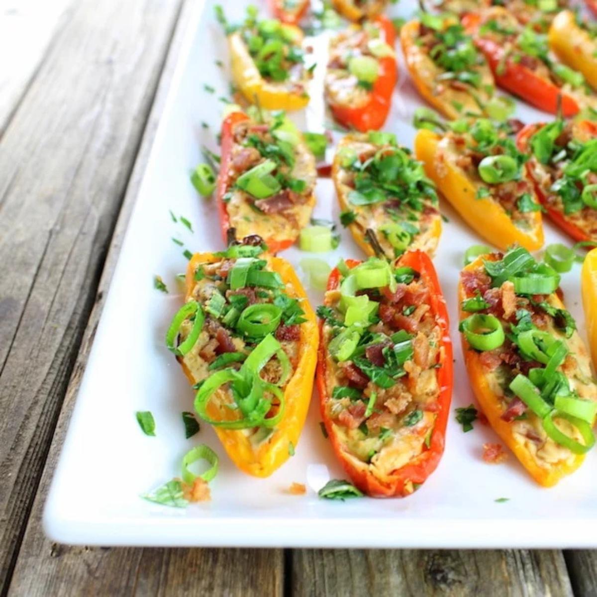 Baked mini peppers stuffed with a creamy cheese mixture, topped with crumbled bacon and chopped green onions. Served on a white platter.