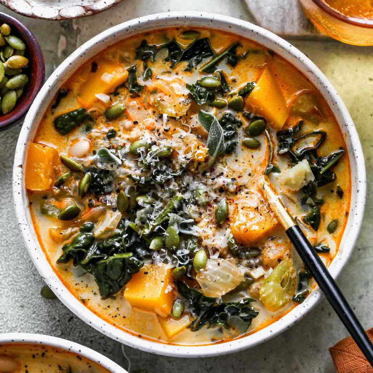 A bowl of creamy butternut squash soup with kale, white beans, and pumpkin seeds. The soup is a rich orange color and is topped with a dollop of sour cream, shaved Parmesan cheese, and fresh sage leaves.