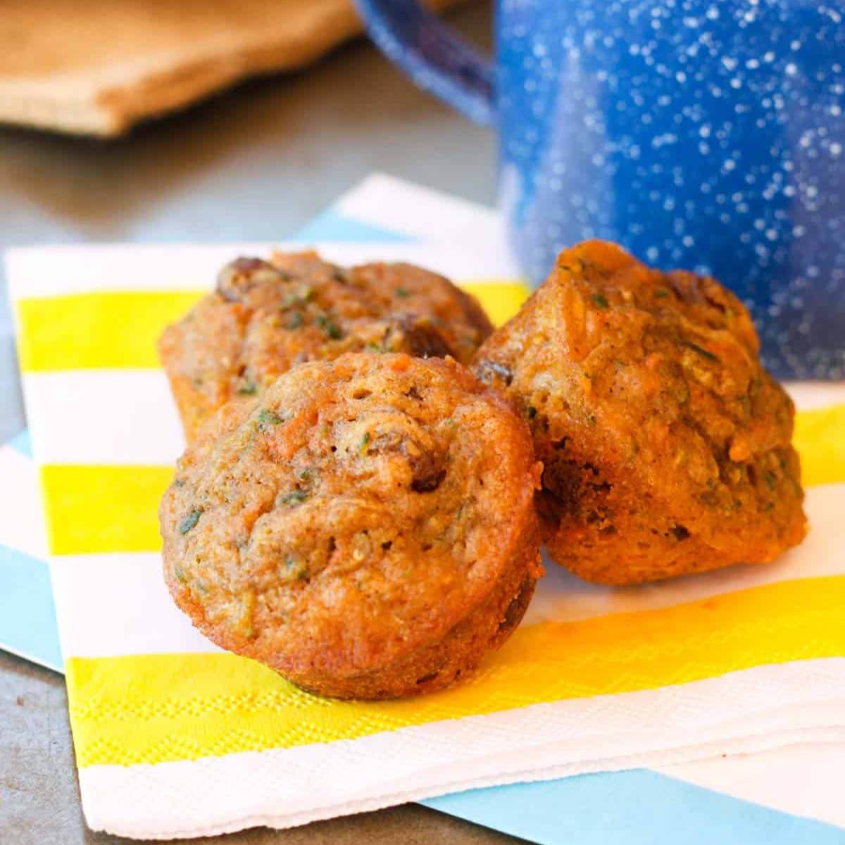 Three mini carrot muffins on a yellow and blue striped napkin, with a blue mug in the background.