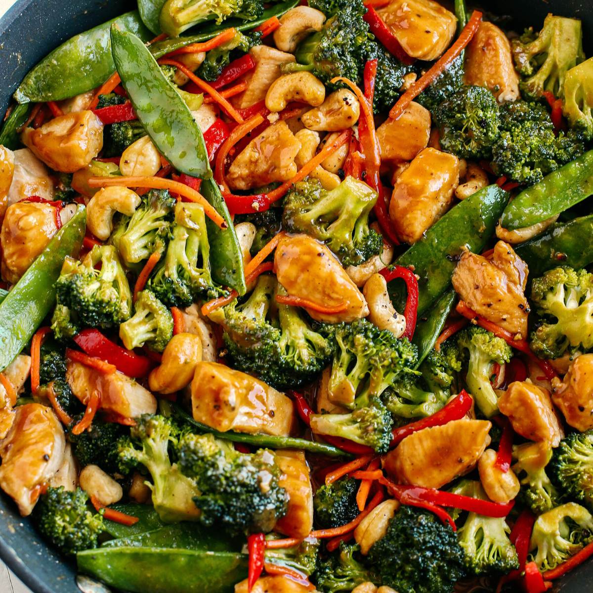  A skillet filled with a colorful stir-fry dish featuring chicken, broccoli, carrots, snow peas, red bell peppers, and cashews. The dish is coated in a glossy sauce.