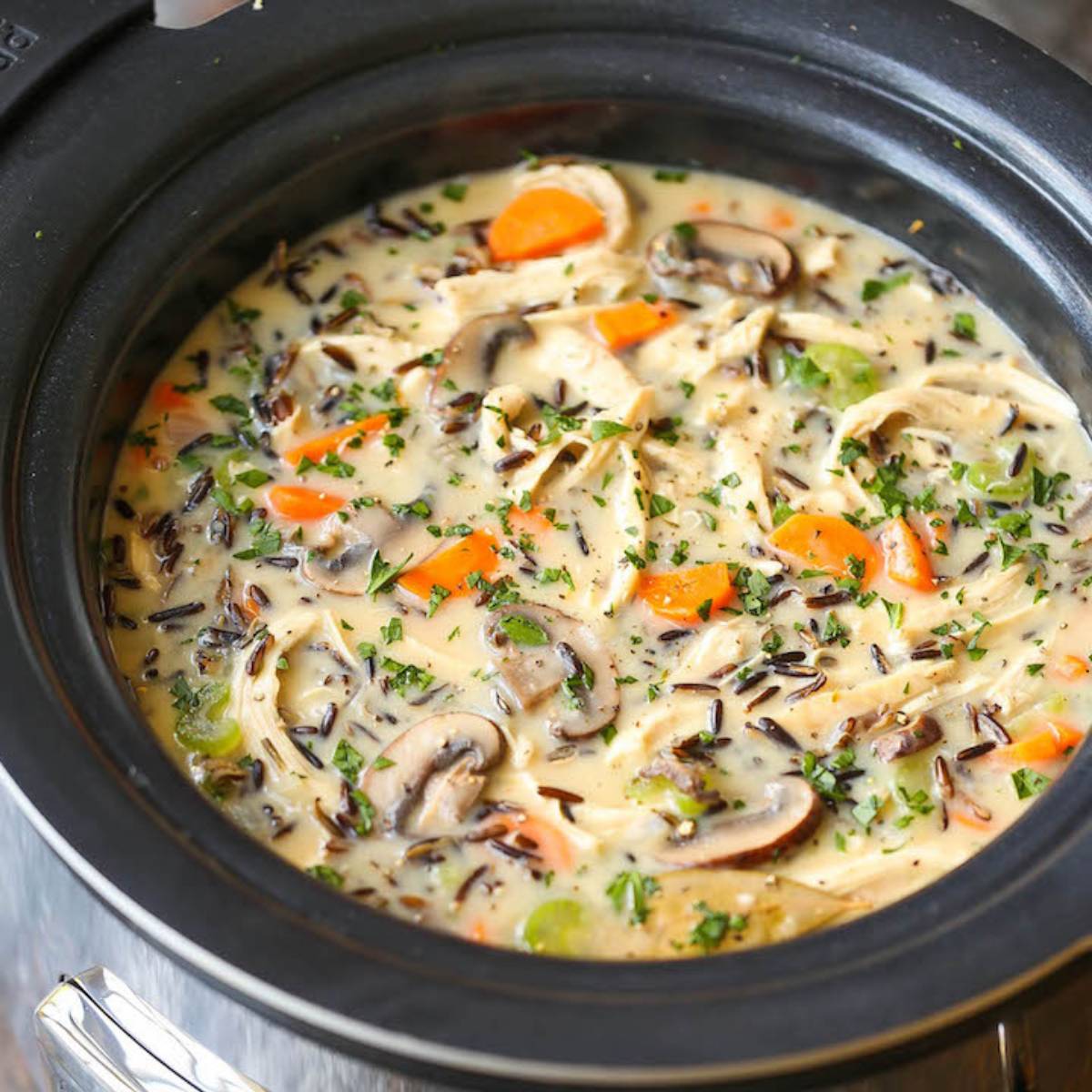 A slow cooker filled with a creamy chicken and wild rice soup. The soup contains shredded chicken, wild rice, carrots, mushrooms, and a creamy broth. Fresh parsley is sprinkled on top.