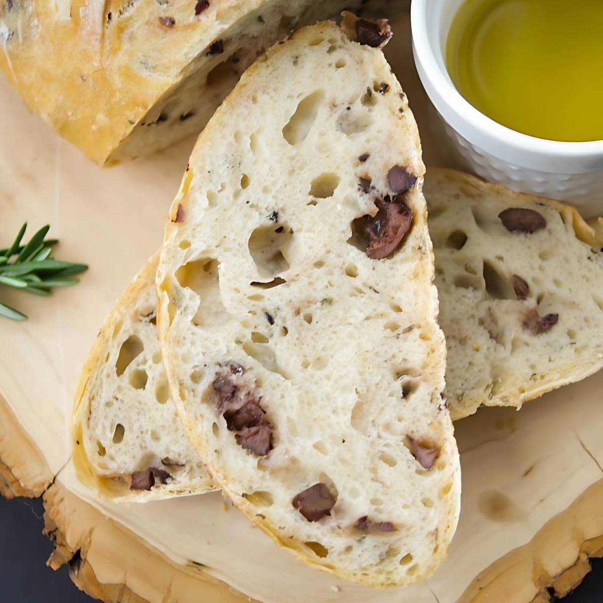 A close-up of slices of crusty bread with black olives embedded throughout. A small bowl of olive oil is visible in the background.