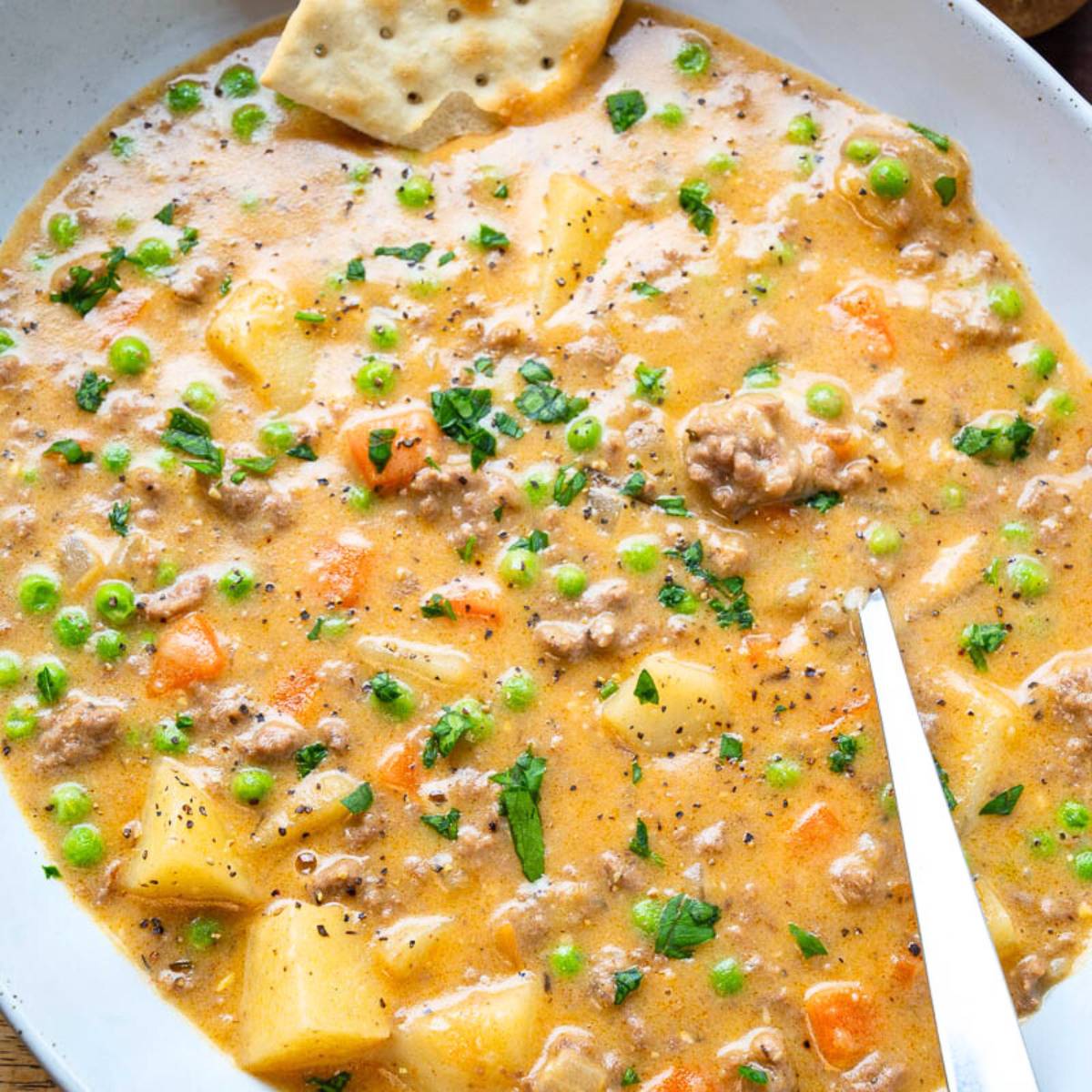 A bowl of creamy shepherd's pie soup, filled with ground beef, potatoes, carrots, peas, and corn. The soup is topped with fresh parsley and a cracker.