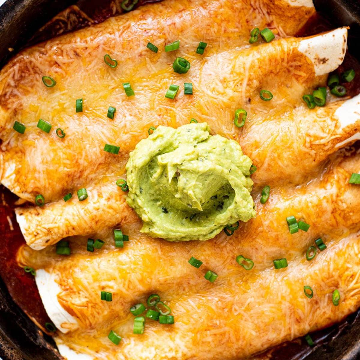 A close-up photo of a skillet filled with chicken enchiladas topped with melted cheese, a dollop of guacamole, and chopped green onions.