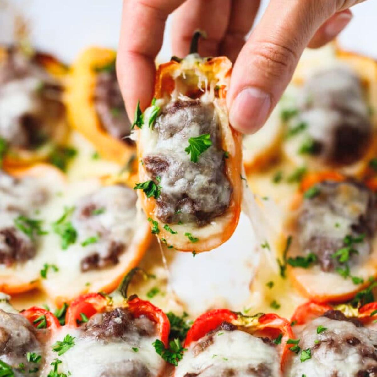  A hand picking up a stuffed mini pepper filled with a meatball and cheese, with melted cheese pulling away.