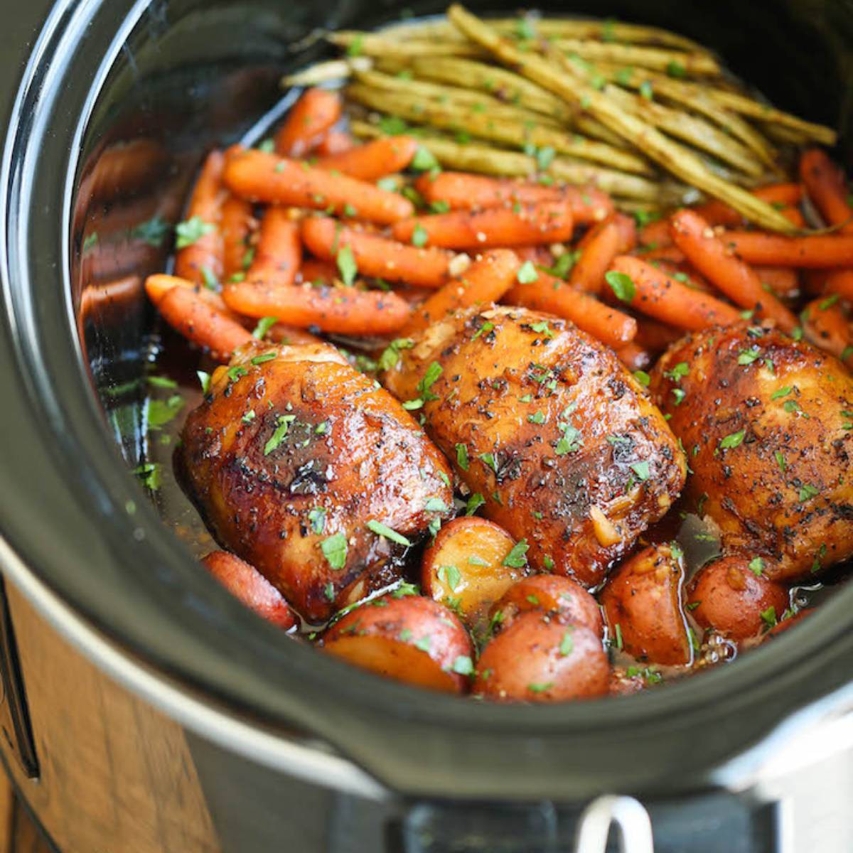 A slow cooker filled with honey garlic chicken and vegetables. The chicken is coated in a sweet and savory sauce, and the vegetables include carrots and baby potatoes. 