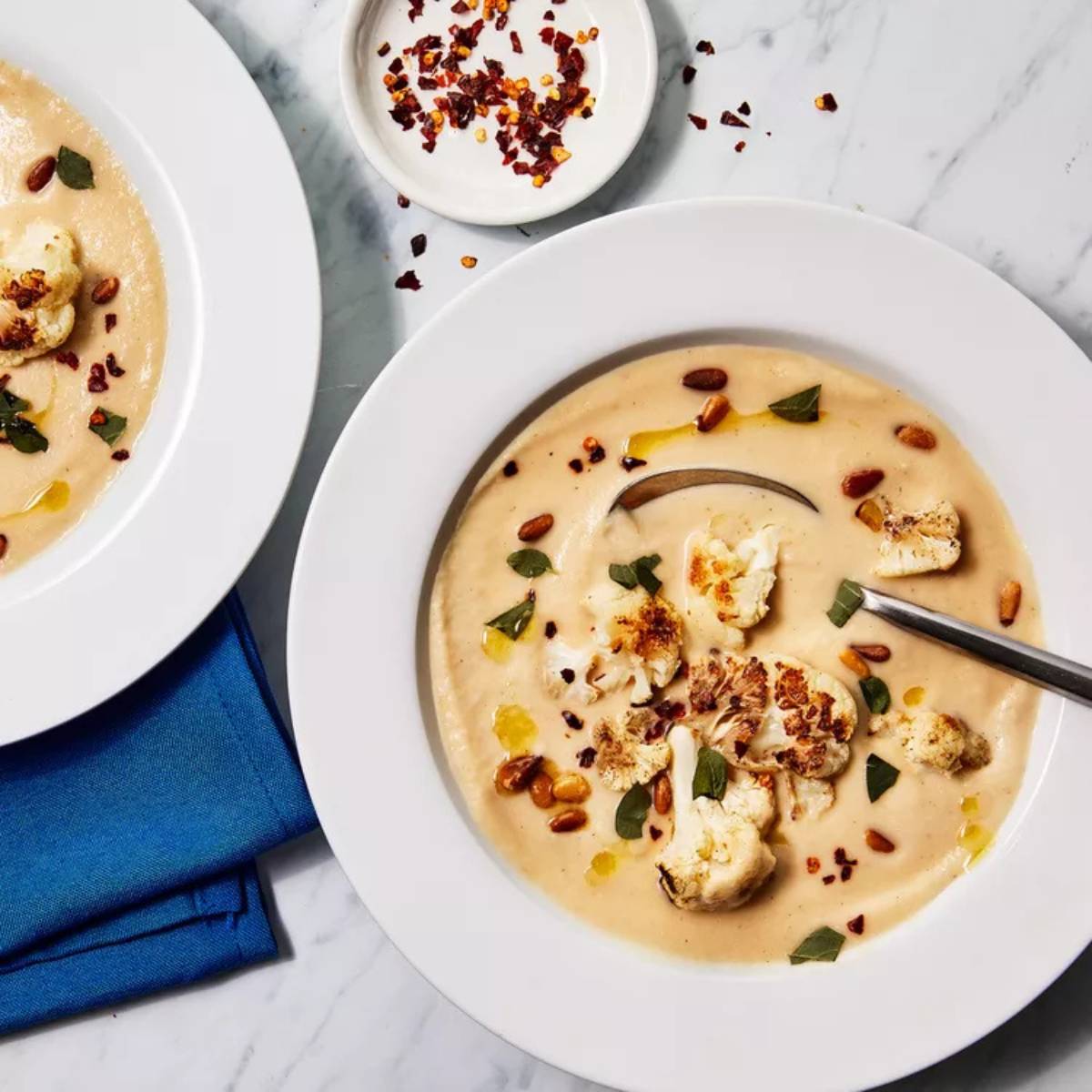 Two white bowls filled with creamy roasted cauliflower soup, topped with roasted cauliflower florets, pine nuts, red pepper flakes, and fresh herbs.