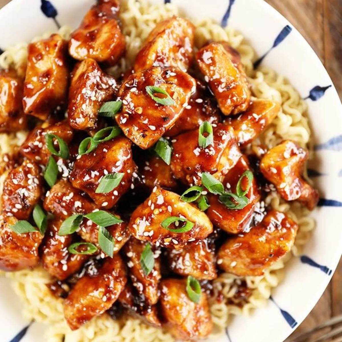  A bowl of General Tso's chicken served over ramen noodles. The chicken is coated in a sweet and spicy sauce and garnished with sesame seeds and chopped green onions.