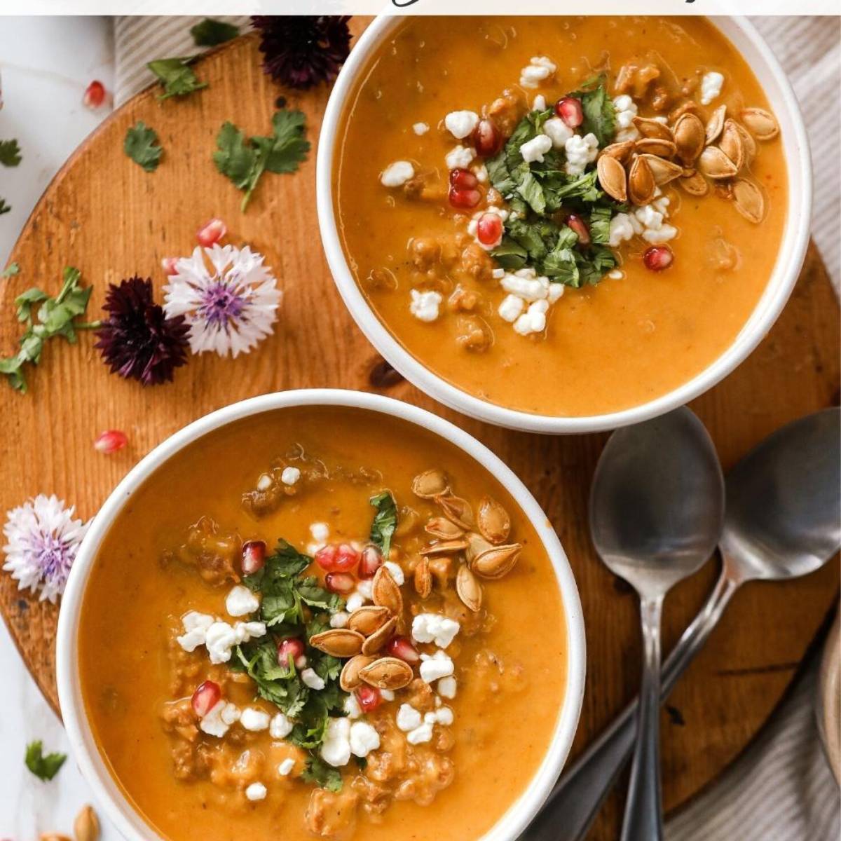 Two bowls of creamy pumpkin chorizo soup, topped with crumbled feta cheese, pomegranate seeds, toasted pumpkin seeds, and fresh cilantro. The soup is a vibrant orange color and is served in white bowls on a wooden board.