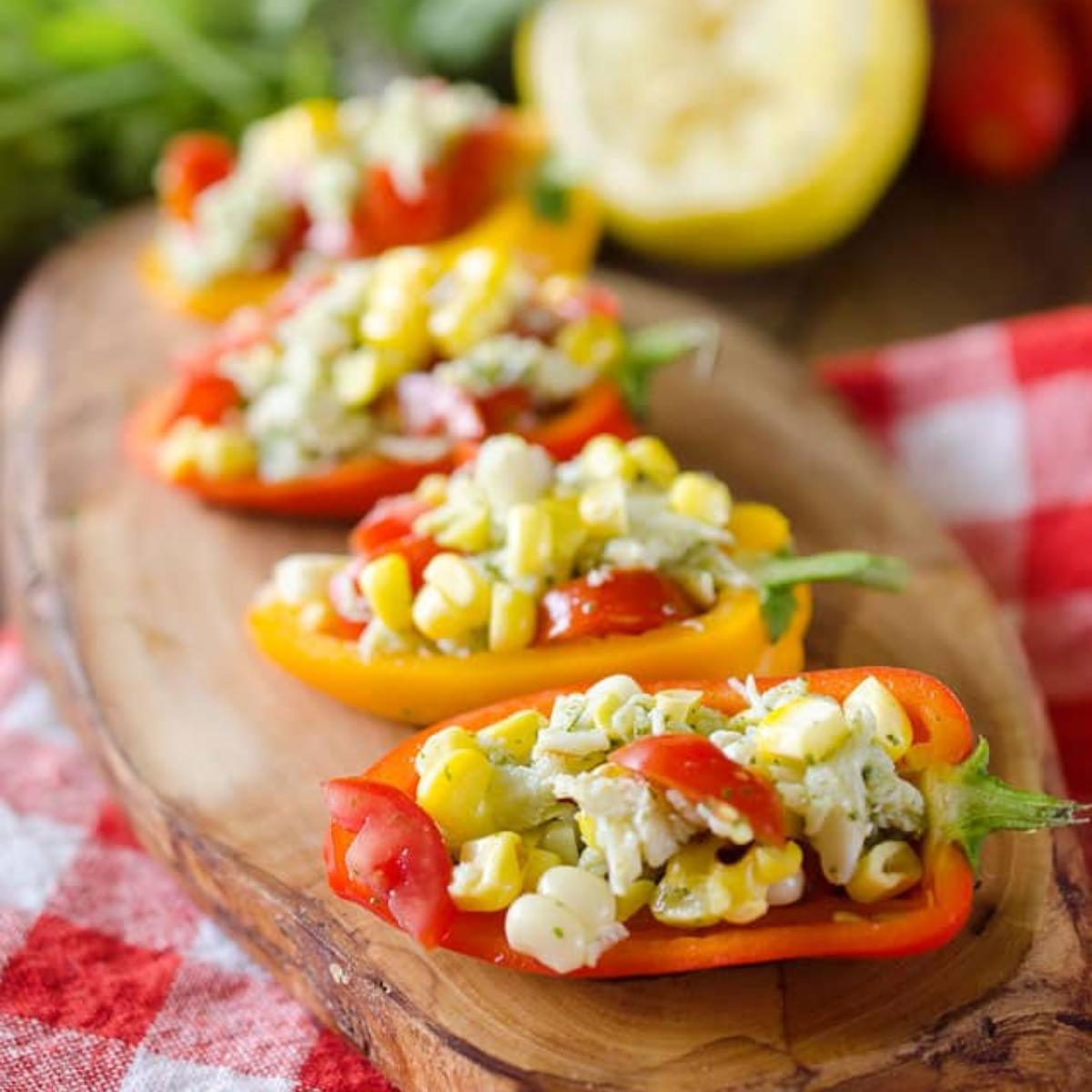 Mini peppers stuffed with a creamy, colorful filling made of cheese, corn, and tomatoes. Served on a wooden board with a lemon wedge in the background.