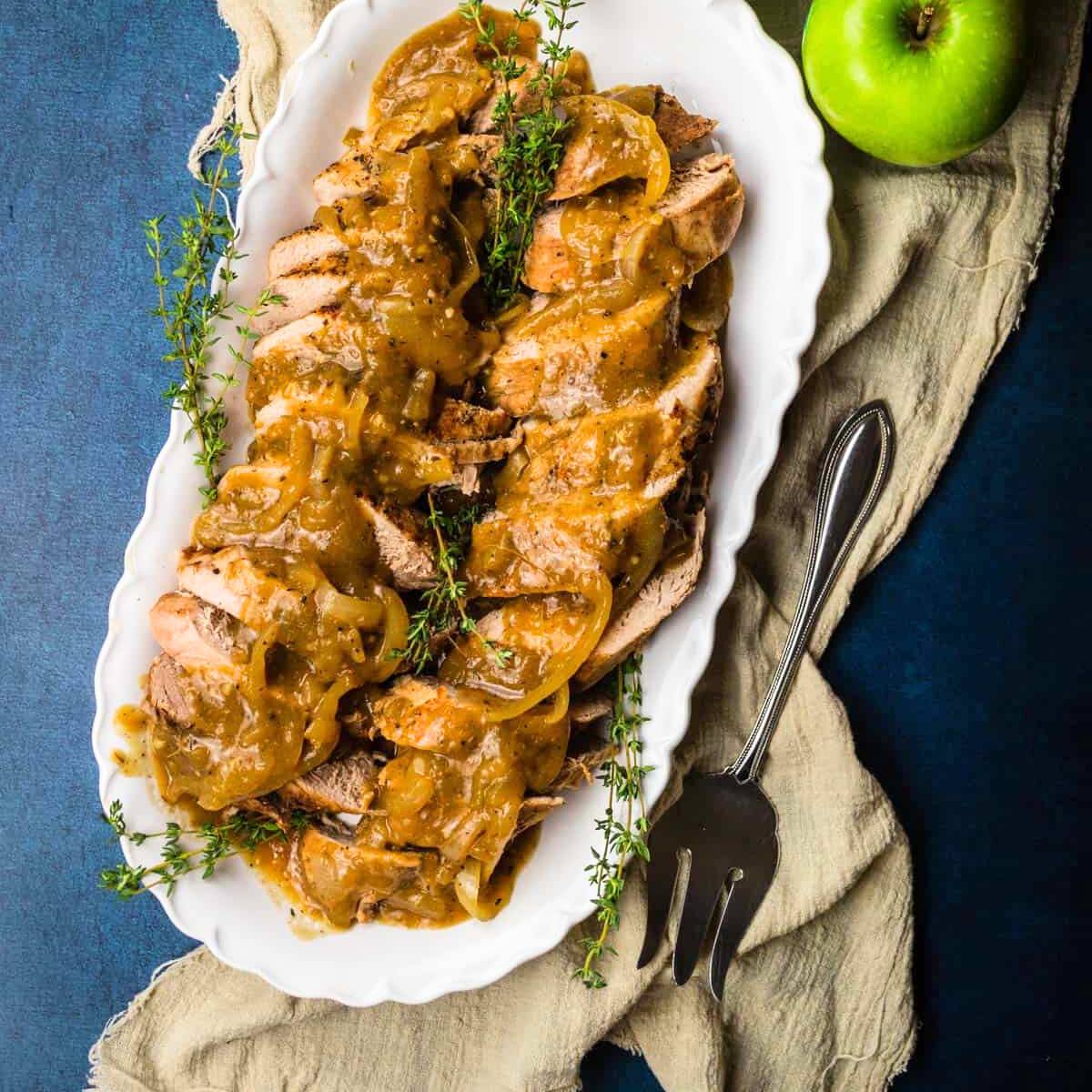 A platter of sliced pork tenderloin served with an apple-onion sauce. Fresh thyme sprigs are garnishing the dish. A whole green apple is visible in the background.