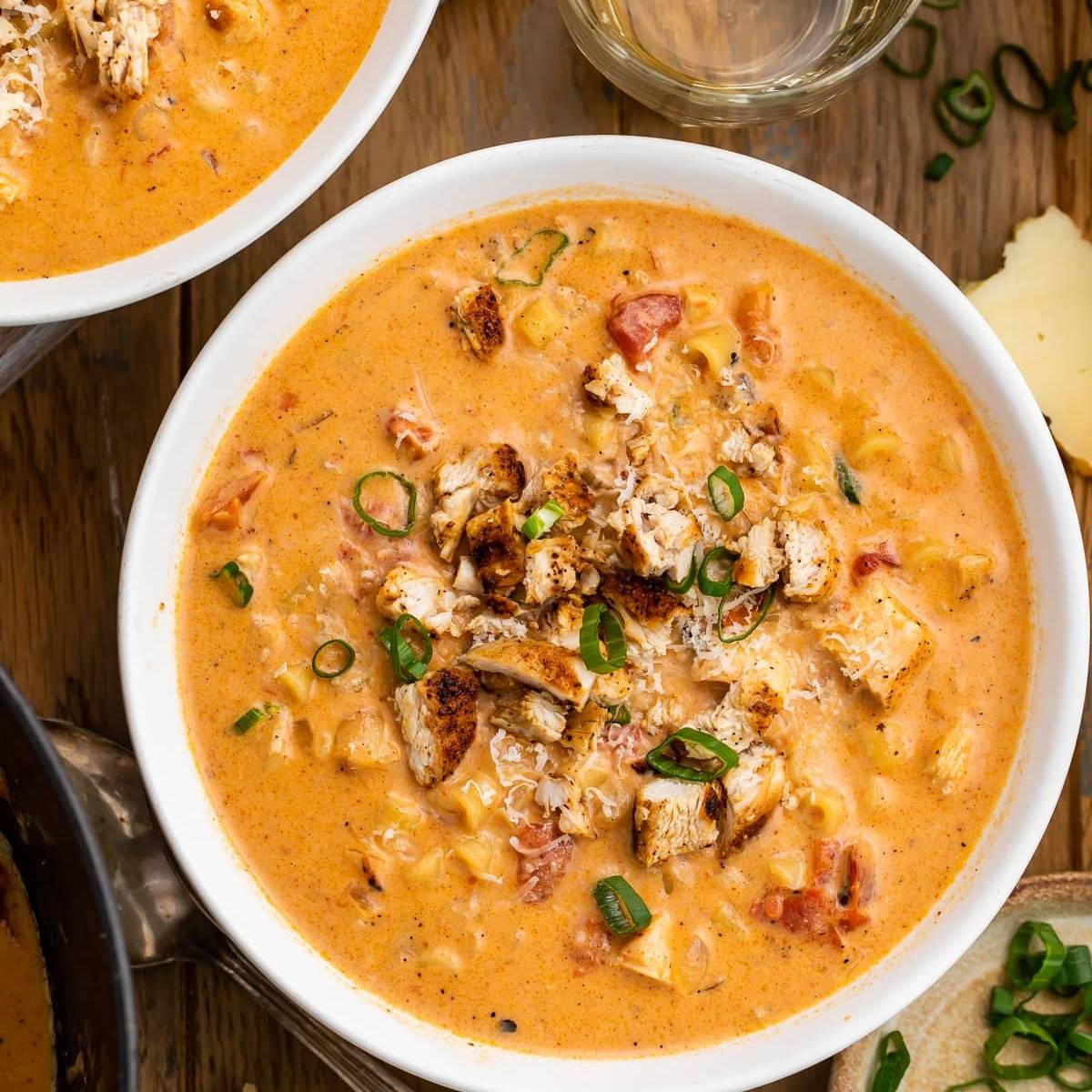 Two bowls of creamy chicken and sausage soup, topped with shredded chicken, green onions, and shredded cheese. The soup is a vibrant orange color and contains pasta and vegetables.