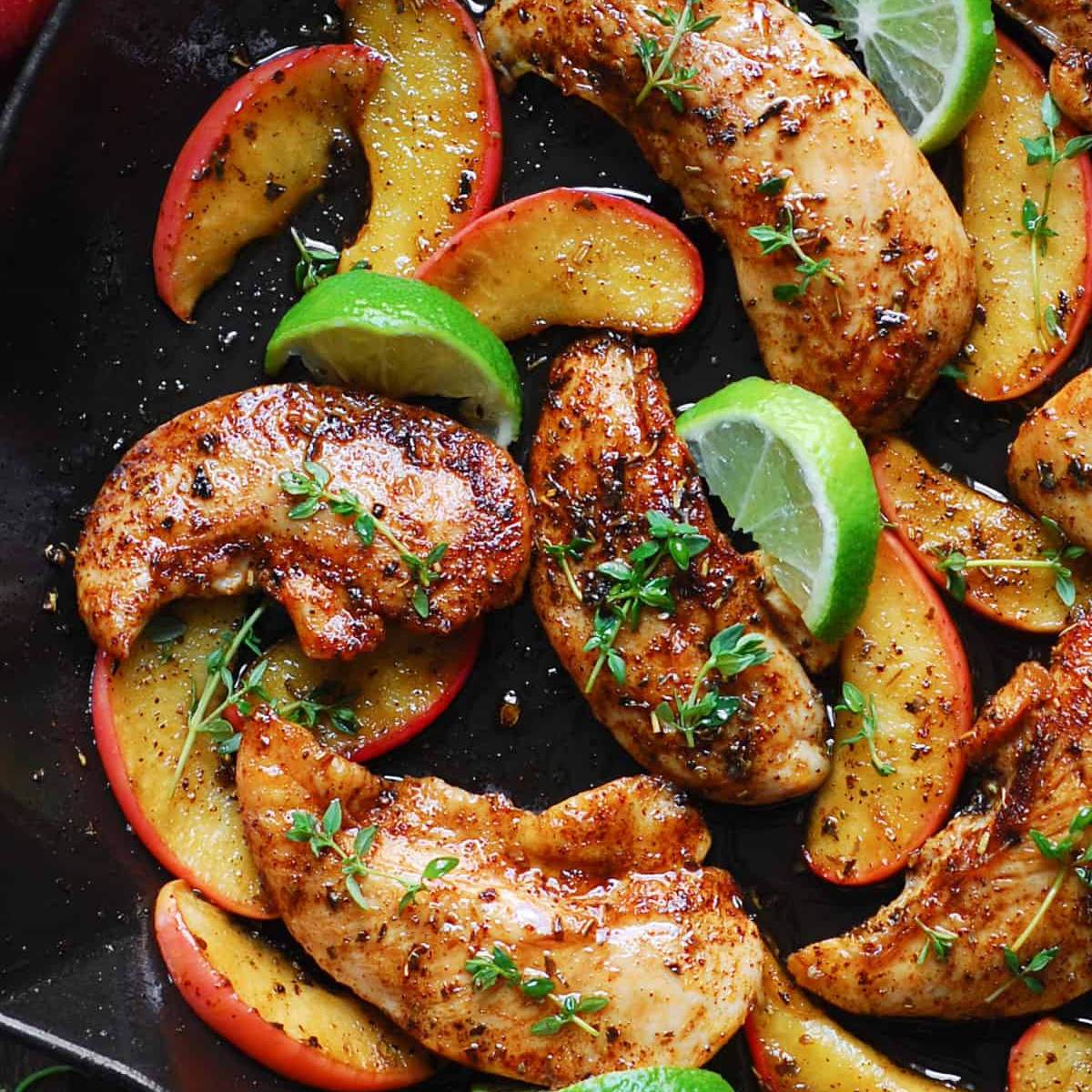 A skillet filled with seasoned chicken and sautéed apple slices. Lime wedges and fresh thyme are sprinkled on top.