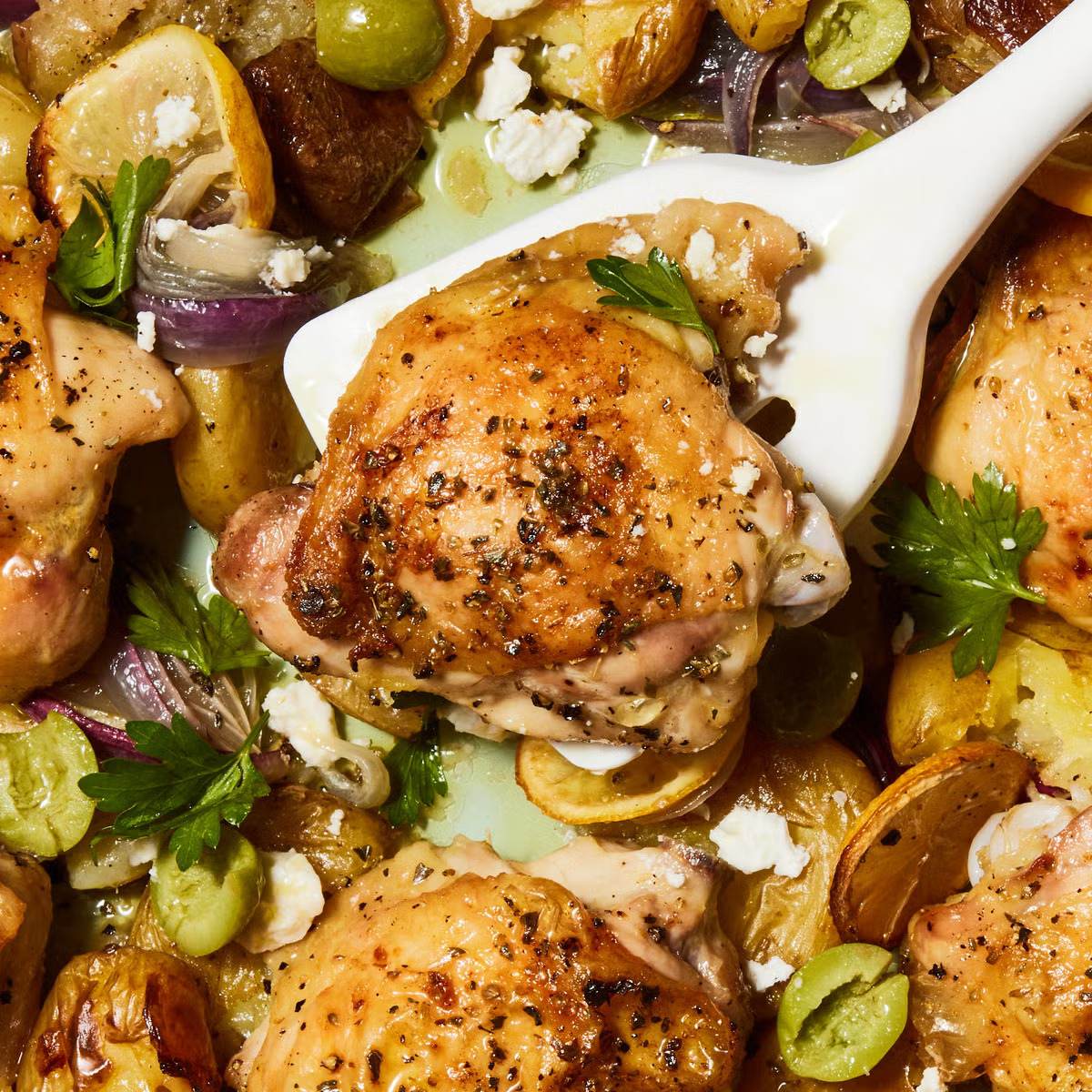 A close-up photo of a sheet pan meal with crispy chicken thighs, roasted potatoes, olives, red onion, lemon wedges, and crumbled feta cheese.